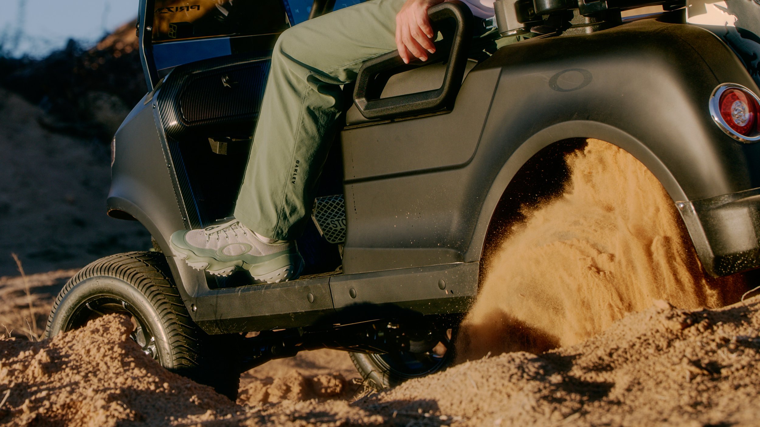 A person driving a small off-road vehicle over sandy terrain, with dust and sand being kicked up by the tires.