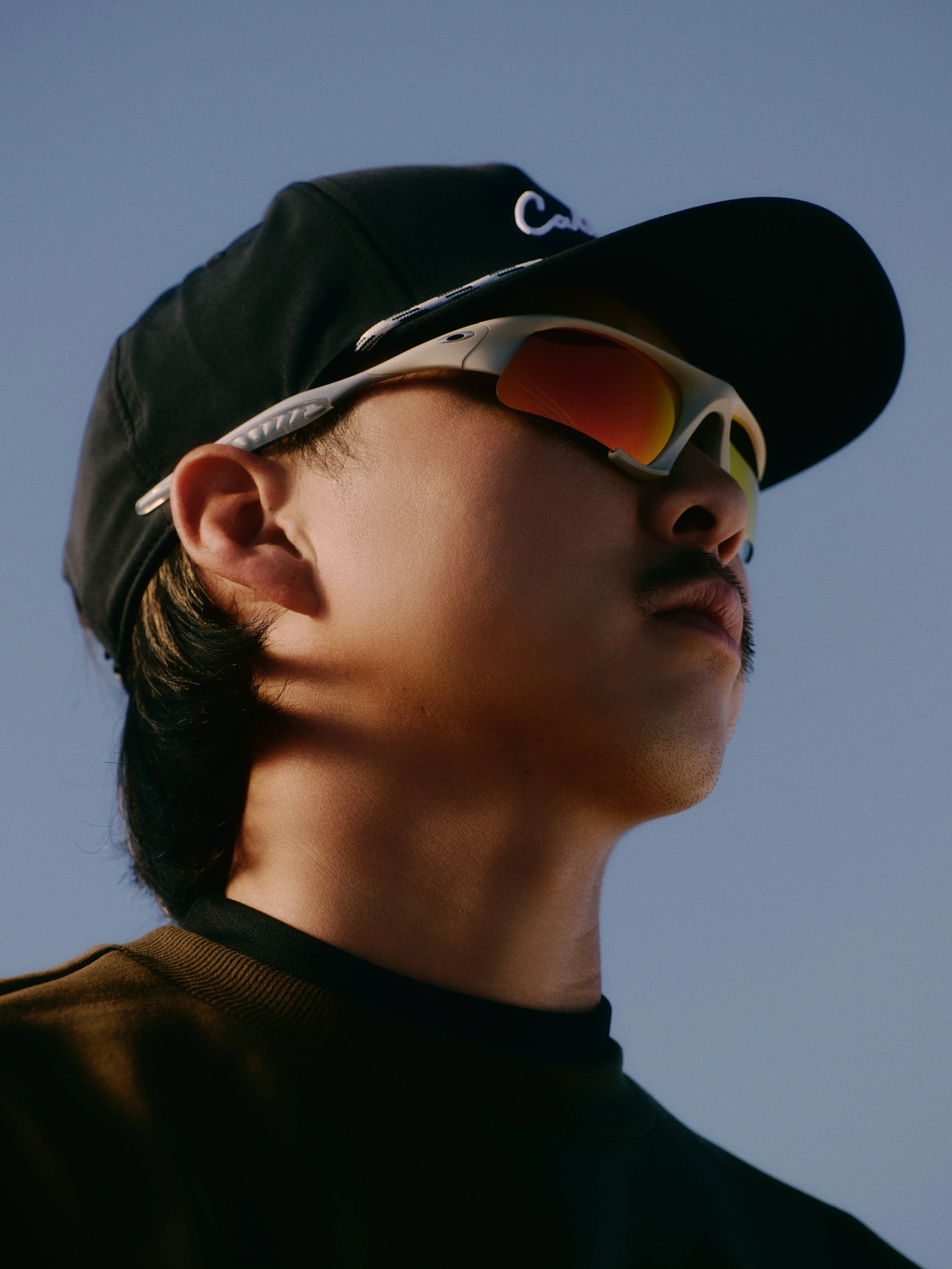 Close-up of a person wearing a black cap with white lettering, white sunglasses with orange lenses, and a black turtleneck, against a clear blue sky.