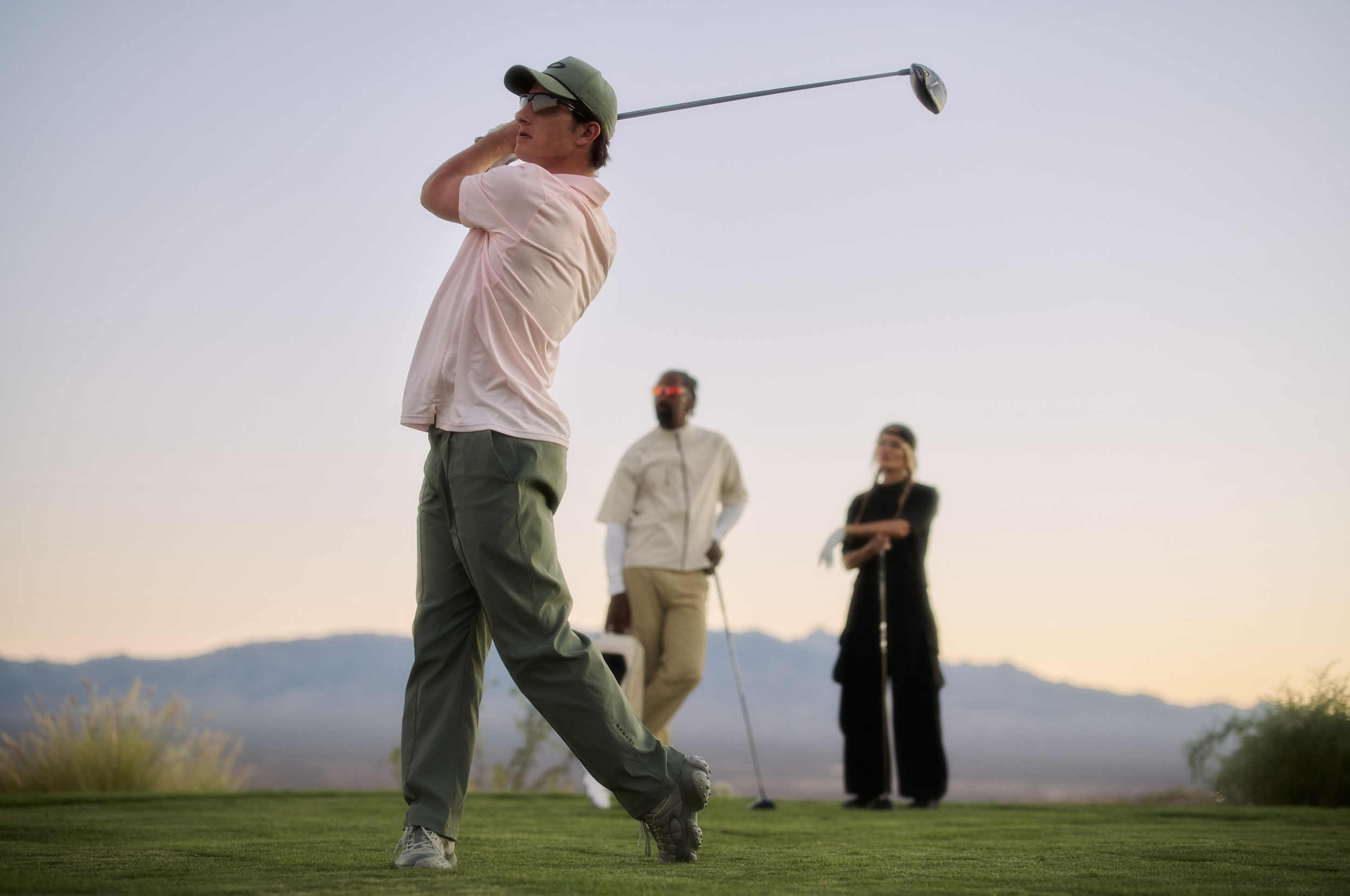 A man in a pink shirt, green pants, and a green cap swings a golf club on a golf course during sunset, with three people in the background watching.