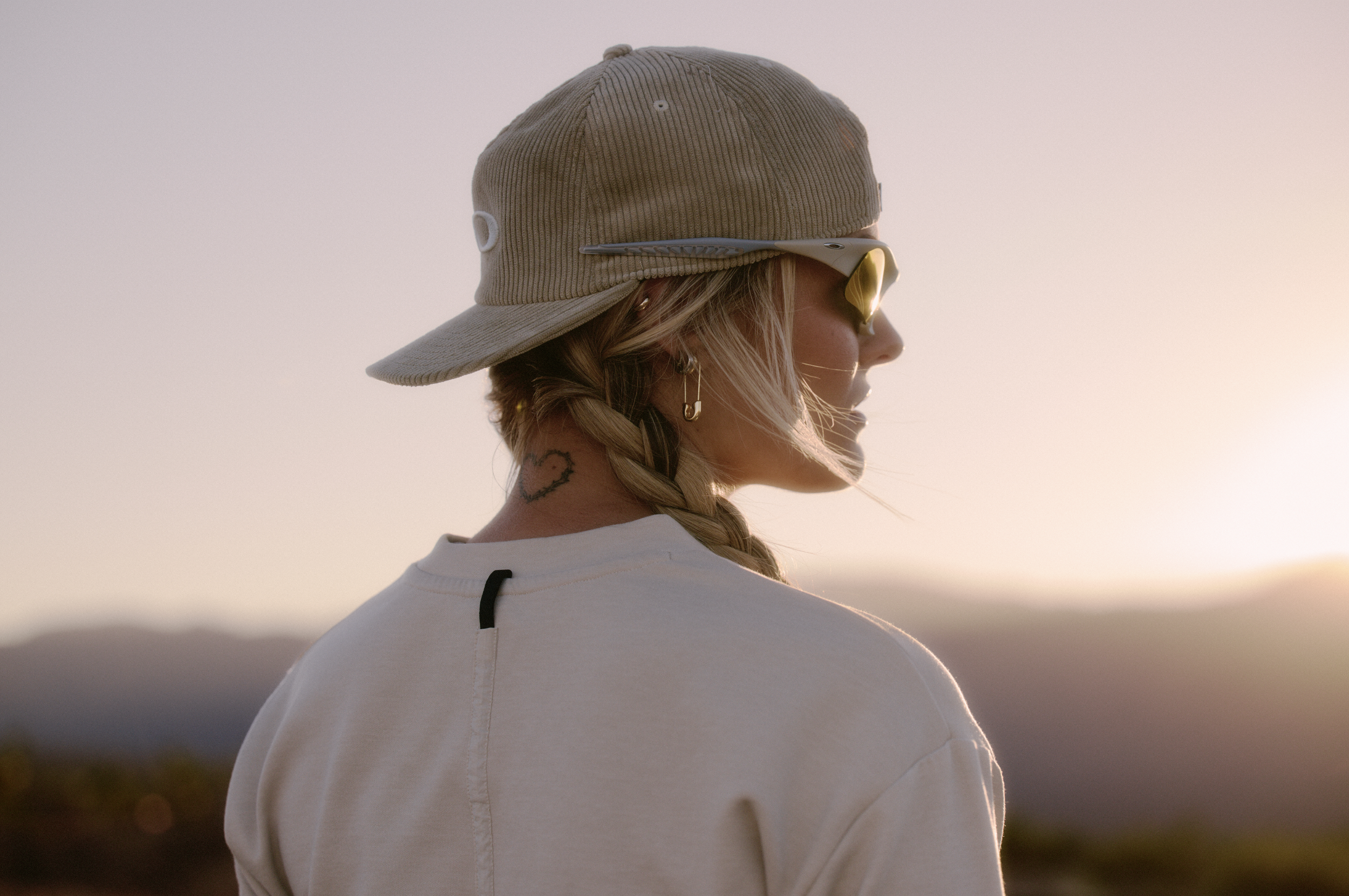 Side profile of a blonde woman wearing a beige hat and yellow sunglasses, standing outdoors at sunset with a blurry natural landscape in the background.