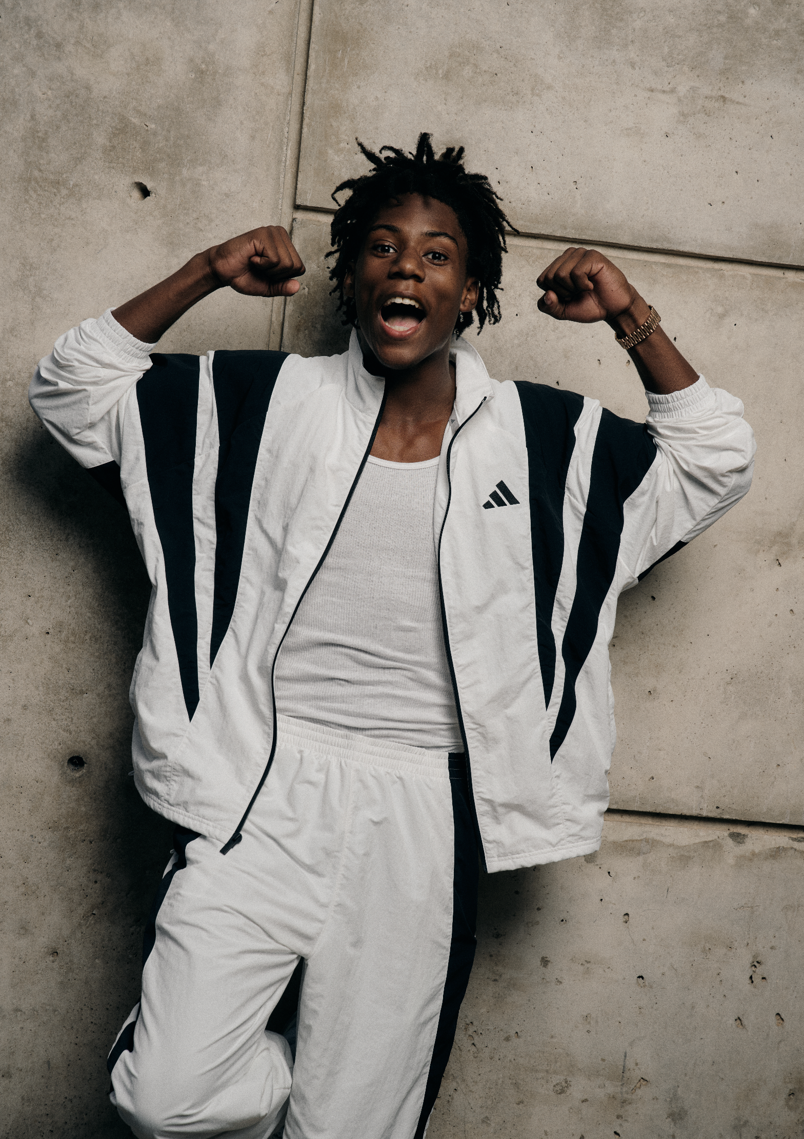 A woman with curly hair is lying on the ground, wearing a white athletic outfit with black stripes, and making a muscle-flexing pose with an open-mouthed expression.