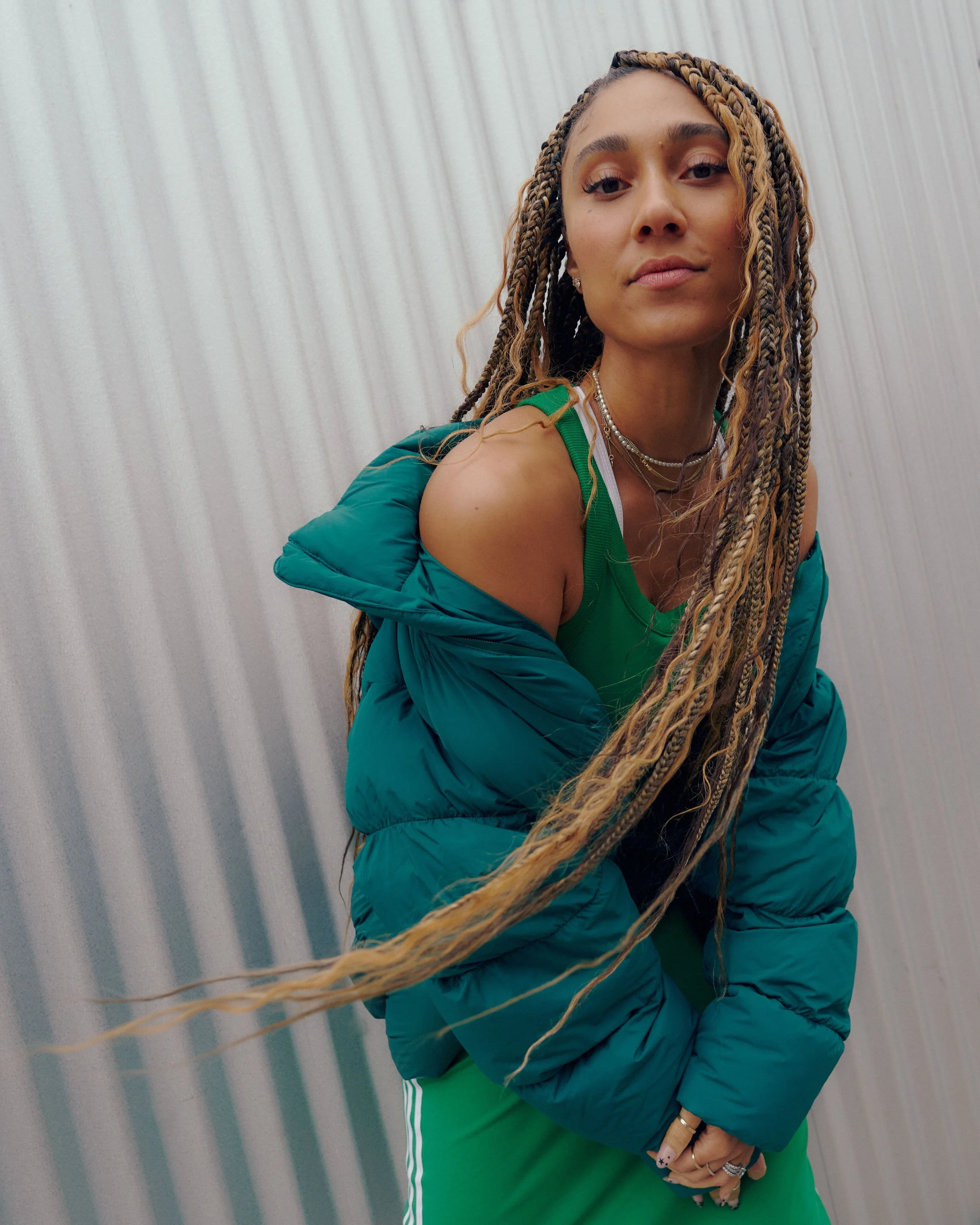A young woman with long blonde braided hair, wearing a green tank top and a teal puffer jacket, standing in front of a textured, white wall. She has rings on her fingers and layered necklaces, with a confident, relaxed expression.