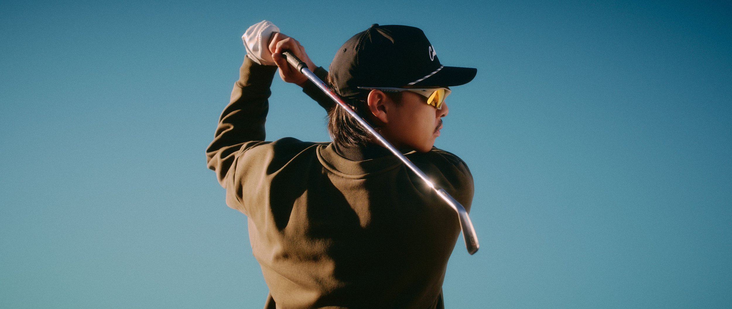 A man wearing a black cap, sunglasses, and a brown jacket, preparing to swing a golf club against a clear blue sky.