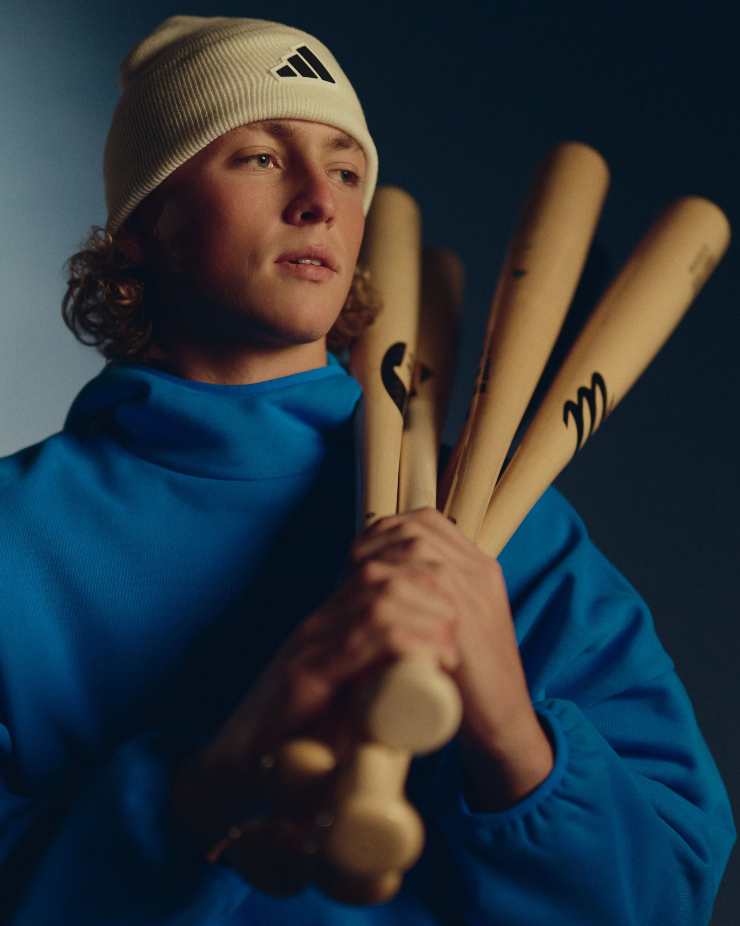 A young man with curly brown hair wearing a white Adidas beanie and a blue jacket, holding four wooden hockey sticks with black branding, against a dark gradient background.