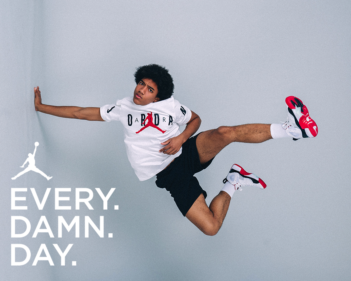A young man in mid-air pose wearing a Nike Jordan shirt, black shorts, and red, white, and black sneakers, against a light grey background with white text that reads, 'EVERY. DAMN. DAY.'