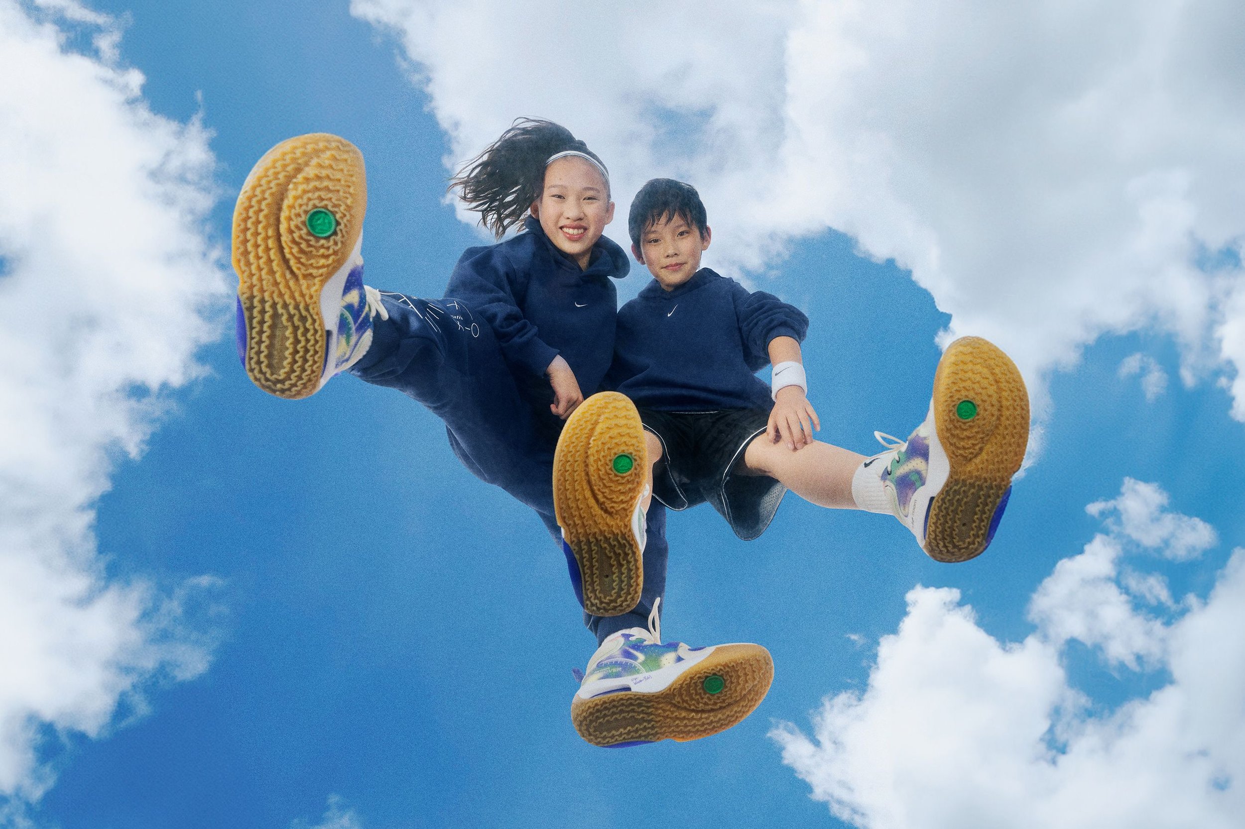 Two kids, a girl and a boy, are seen from below, sitting on a skateboard high in the air against a blue sky with some clouds. The girl has long hair tied back and is smiling. They are wearing dark hoodies, and sneakers.