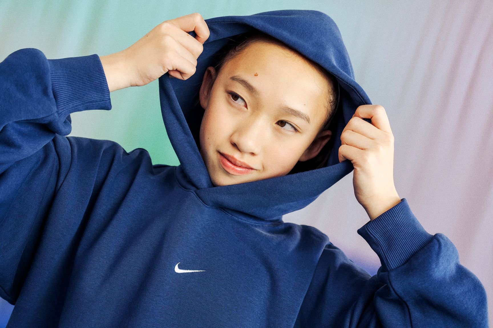 Young person with short hair wearing a blue Nike hoodie, adjusting the hood against a multicolored background.