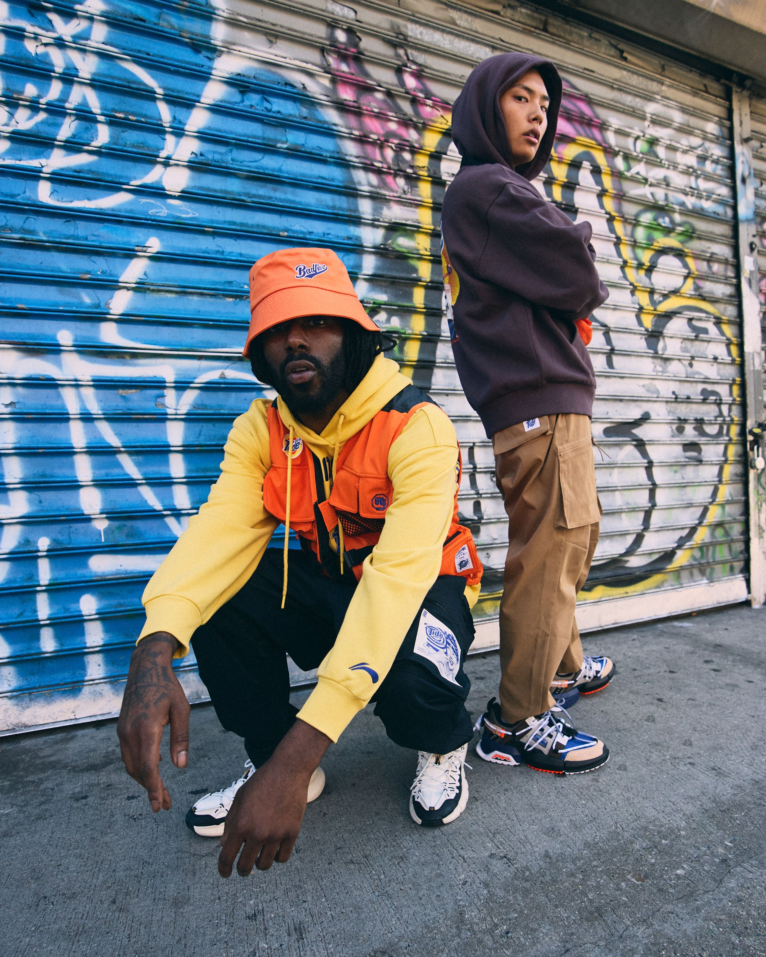 Two young men in streetwear pose in front of a graffiti-covered metal shutter. One is sitting, wearing a yellow hoodie, orange vest, and an orange bucket hat, with dreadlocks. The other stands with his arms crossed, wearing a black hoodie and khaki c