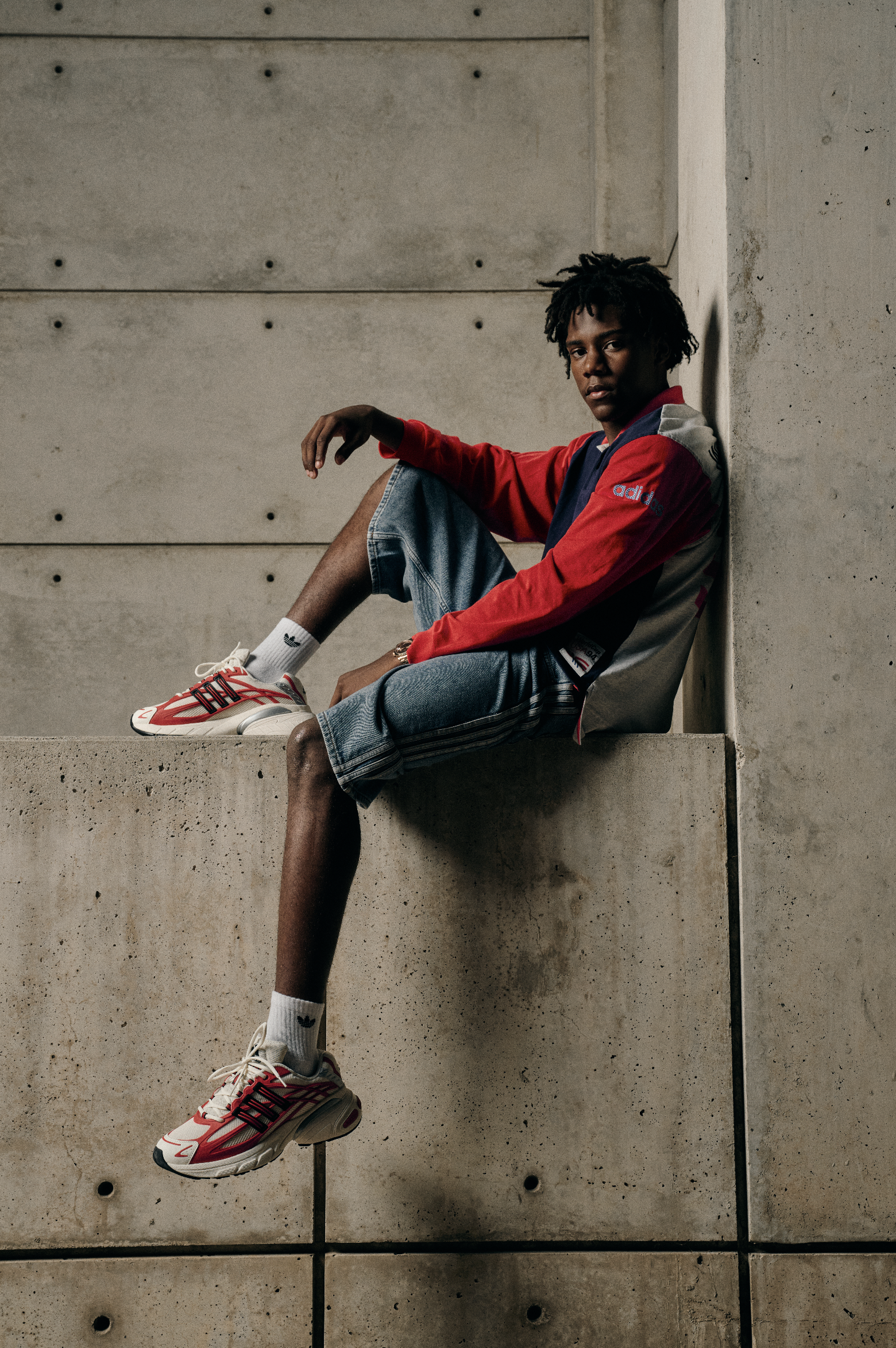 A young man in a red, navy, and beige jacket, denim shorts, white socks, and white sneakers with red and black accents, sitting on a concrete ledge against a concrete wall, with his leg extended and foot resting on the wall.