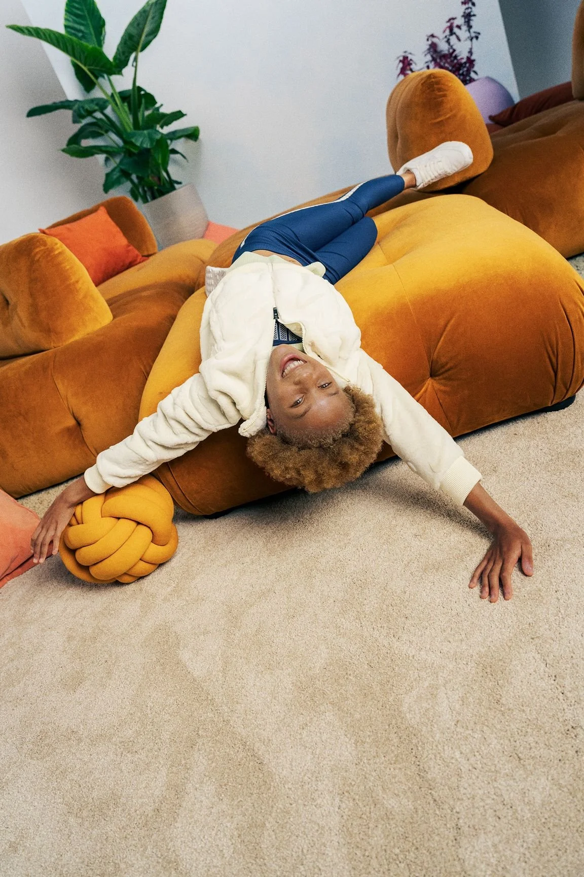 A person with curly hair lying on a mustard-colored sofa, smiling at the camera, with arms and legs stretched out, surrounded by colorful pillows and a large decorative knot pillow, in a living room with a large plant and artwork in the background.