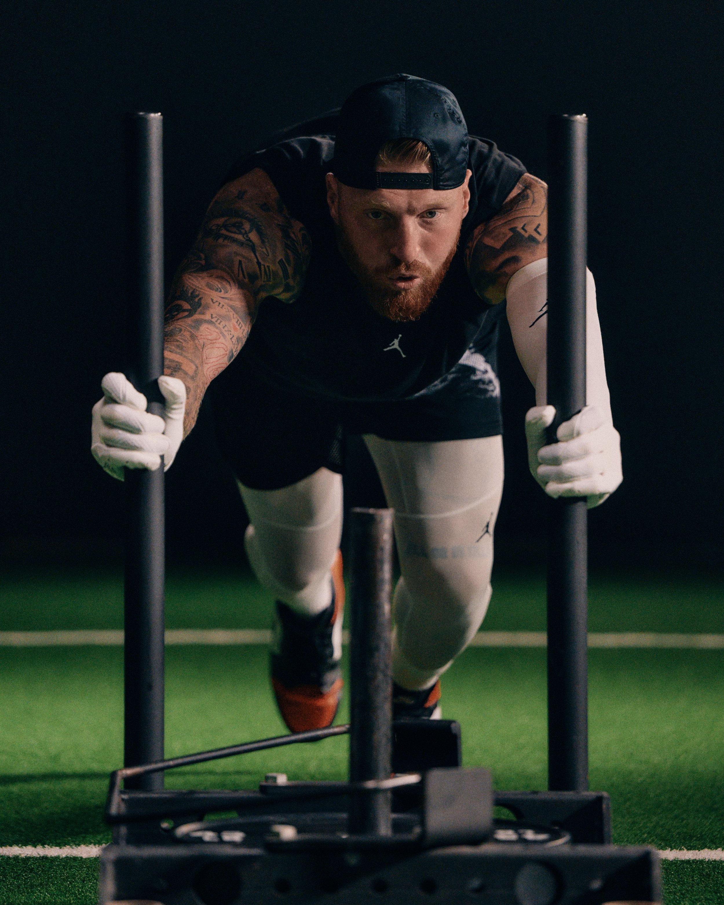 Man with tattoos wearing a cap and athletic gear pushing a sled on a green indoor turf