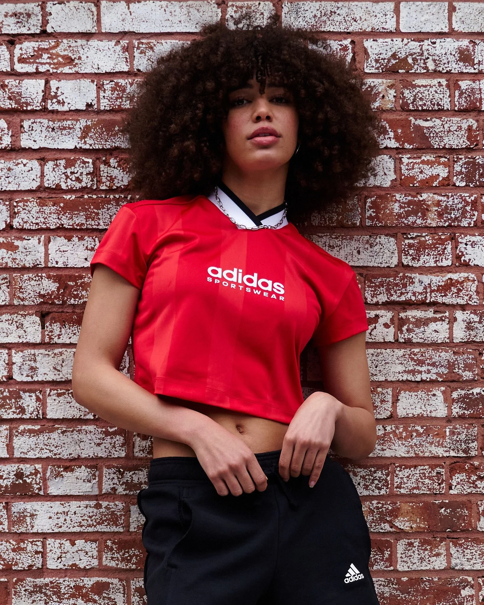 A woman with curly hair wearing a red Adidas sportswear shirt and black Adidas shorts standing against a painted brick wall.