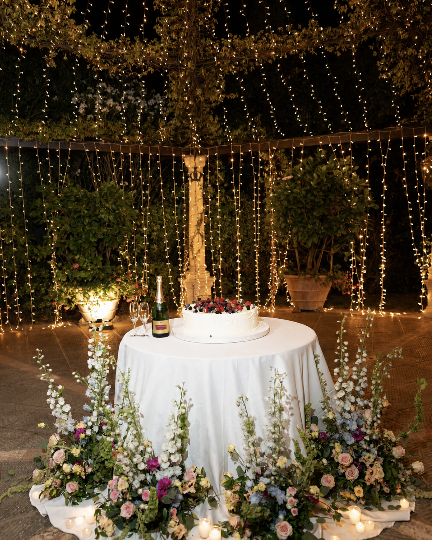 Romantic outdoor wedding cake cutting with candles, flowers, and string lights. Wedding venue is in Tuscany. Cake is done by Imola Catering