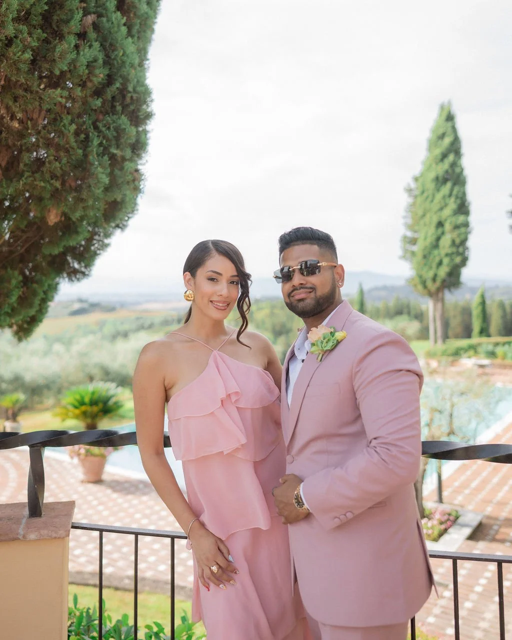 American couple dressed in pink formal attire standing outside on a balcony with trees and a scenic green landscape in the background at an outdoor wedding venue in Florence