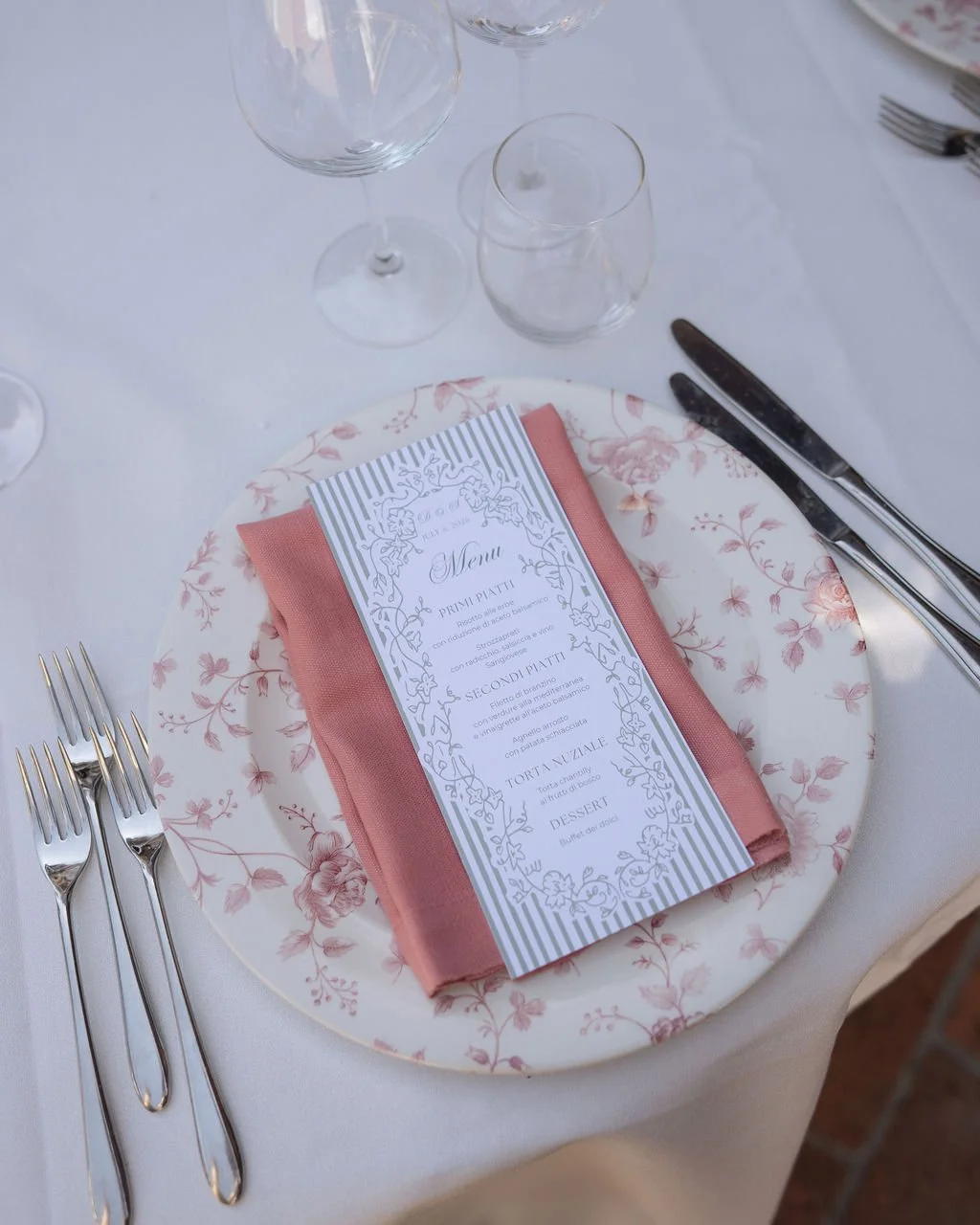 Elegant place setting with floral-patterned plate, pink cloth napkin, menu card on top of napkin, multiple forks on the left, knife and spoon on the right, and empty glasses above.