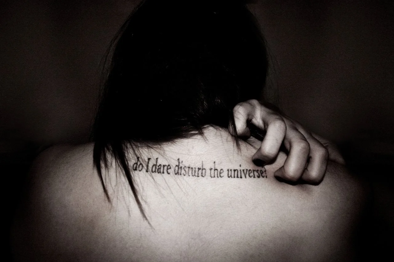 Back of a person with dark hair, showing a tattoo on the upper back that says "Do I dare disturb the universe!" with their hand resting on their shoulder.