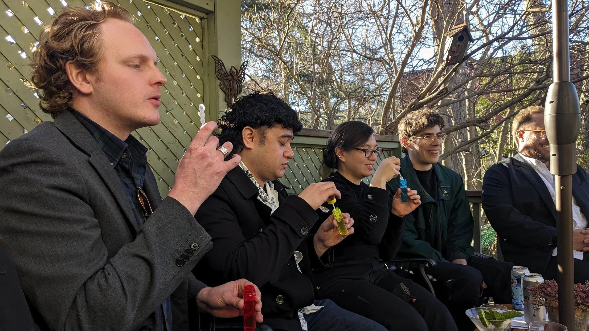 Group of five people sitting outdoors at a table with trees in the background, holding colorful spray bottles, smiling, and enjoying a social gathering.