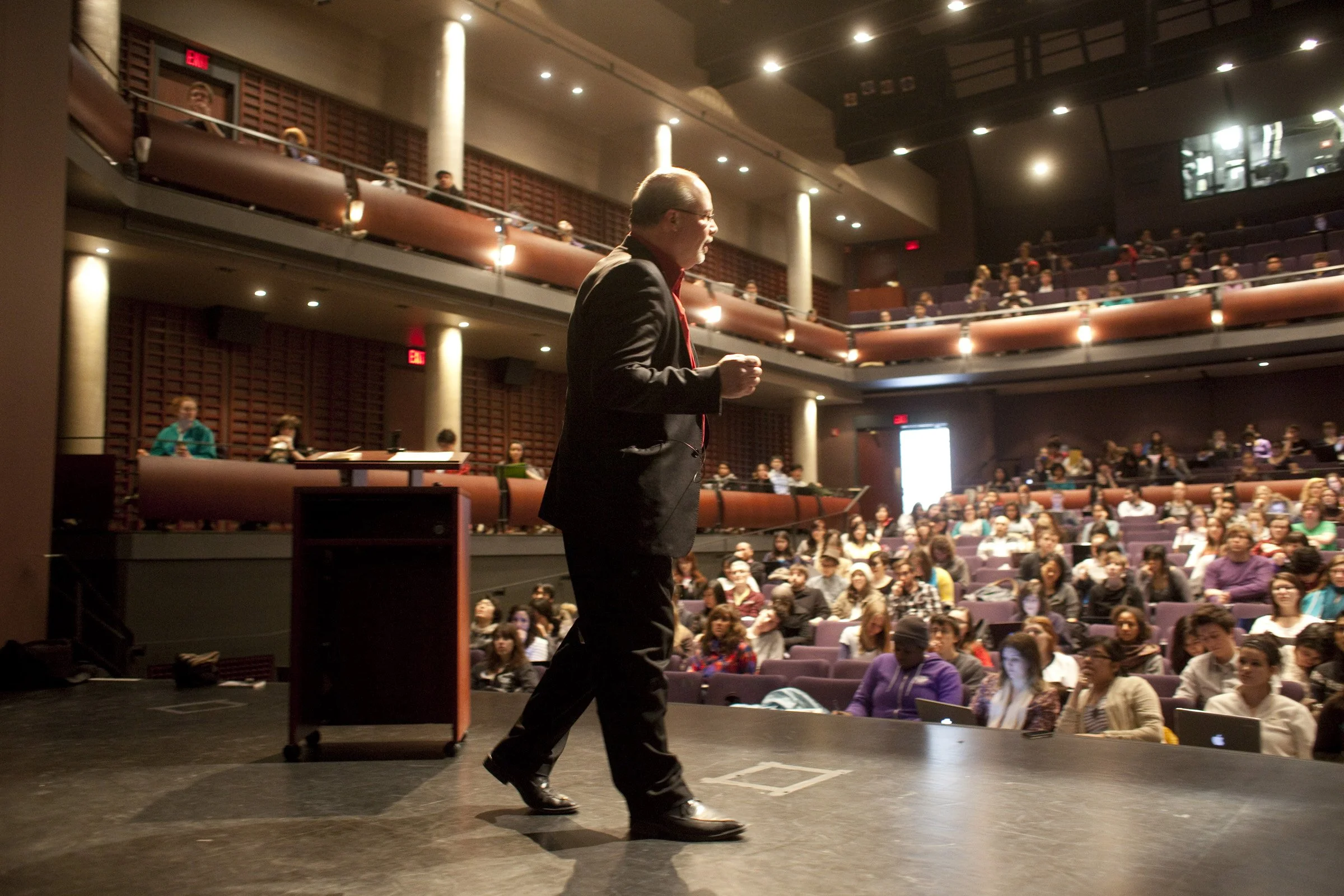 A man in a suit and red shirt speaking on a stage with an audience seated in a theater or auditorium.
