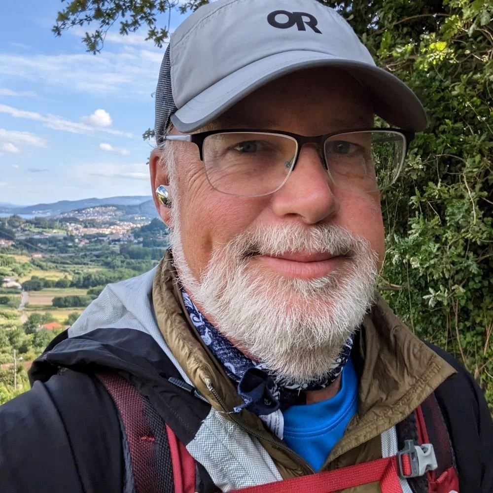 Close-up of a smiling man with white beard and glasses outdoors, wearing a gray cap, outdoor gear, and a backpack, with a scenic landscape of hills and fields in the background.
