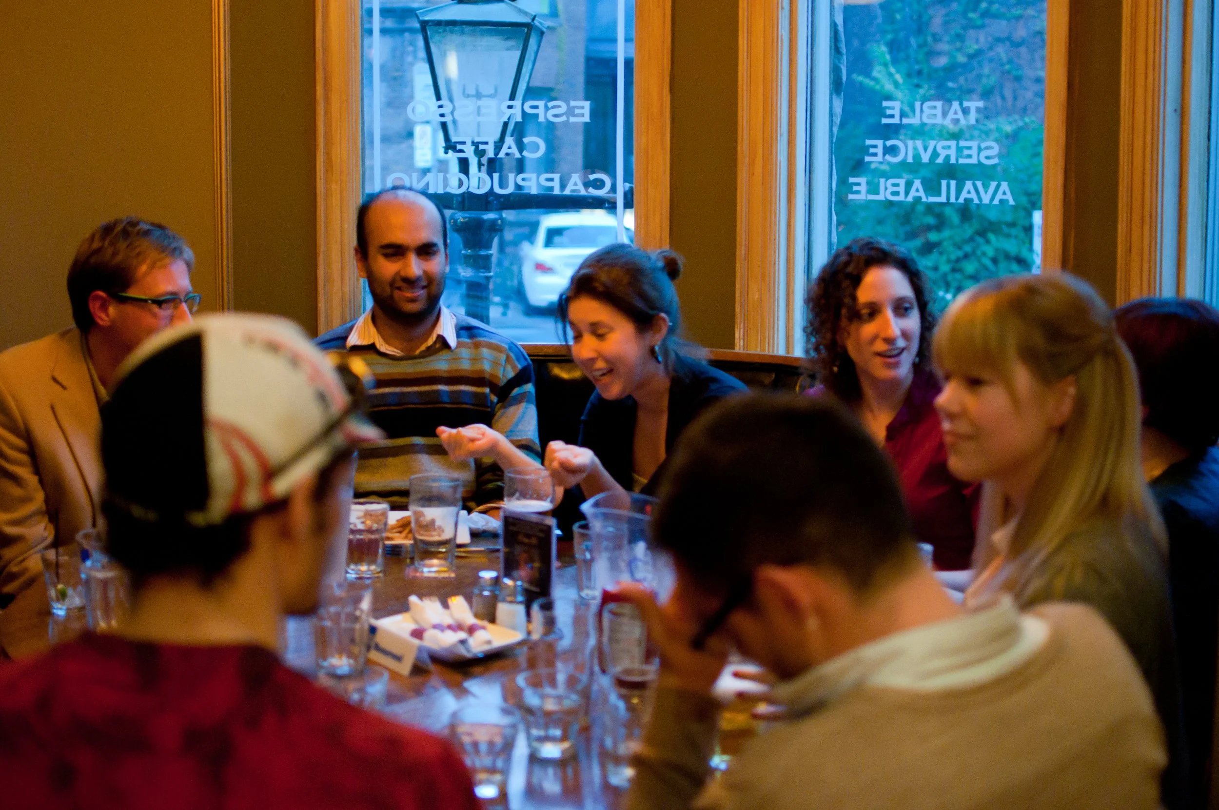A group of people sitting around a table in a restaurant, enjoying conversation and drinks, with windows showing a street scene outside.