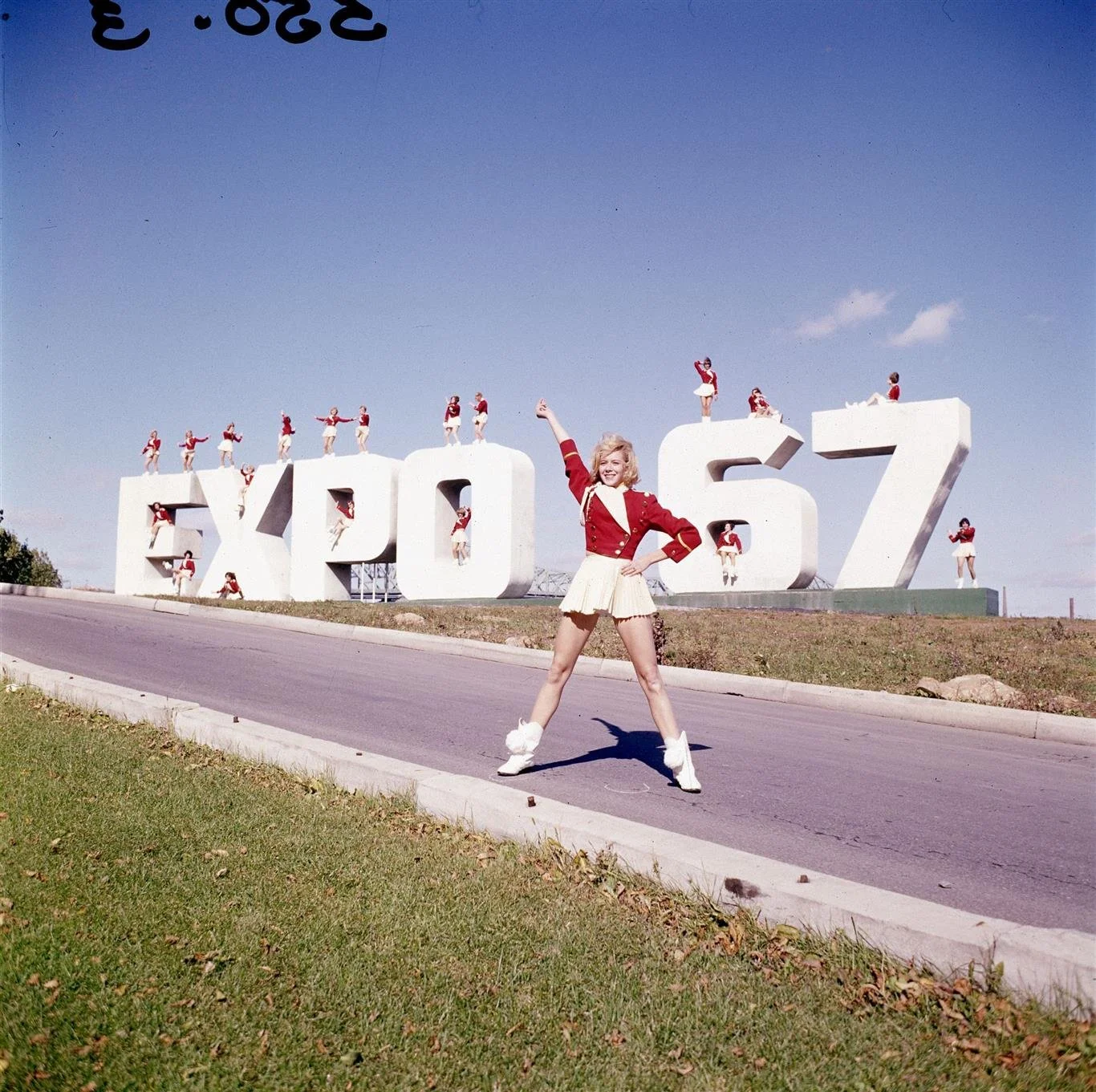 A woman in a cheerleading outfit with a red jacket and white skirt, standing confidently in front of the white 'EXPO 67' sign with smaller women in similar outfits standing on and around the large letters, under a clear blue sky.