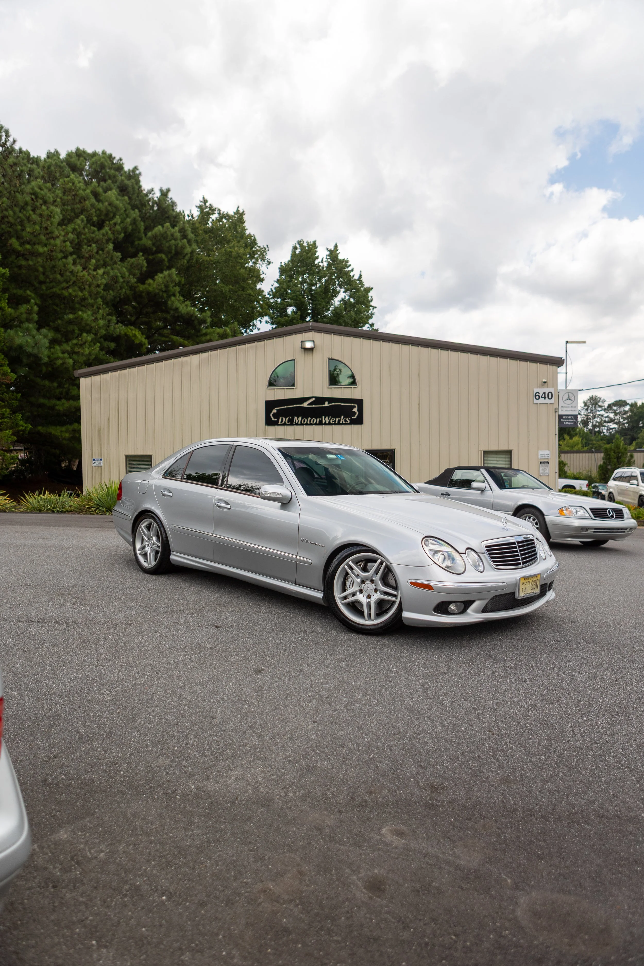 Parking lot in front of a building labeled DC Motor Werks, with a silver Mercedes-Benz car in the foreground and other vehicles parked nearby under cloudy skies.