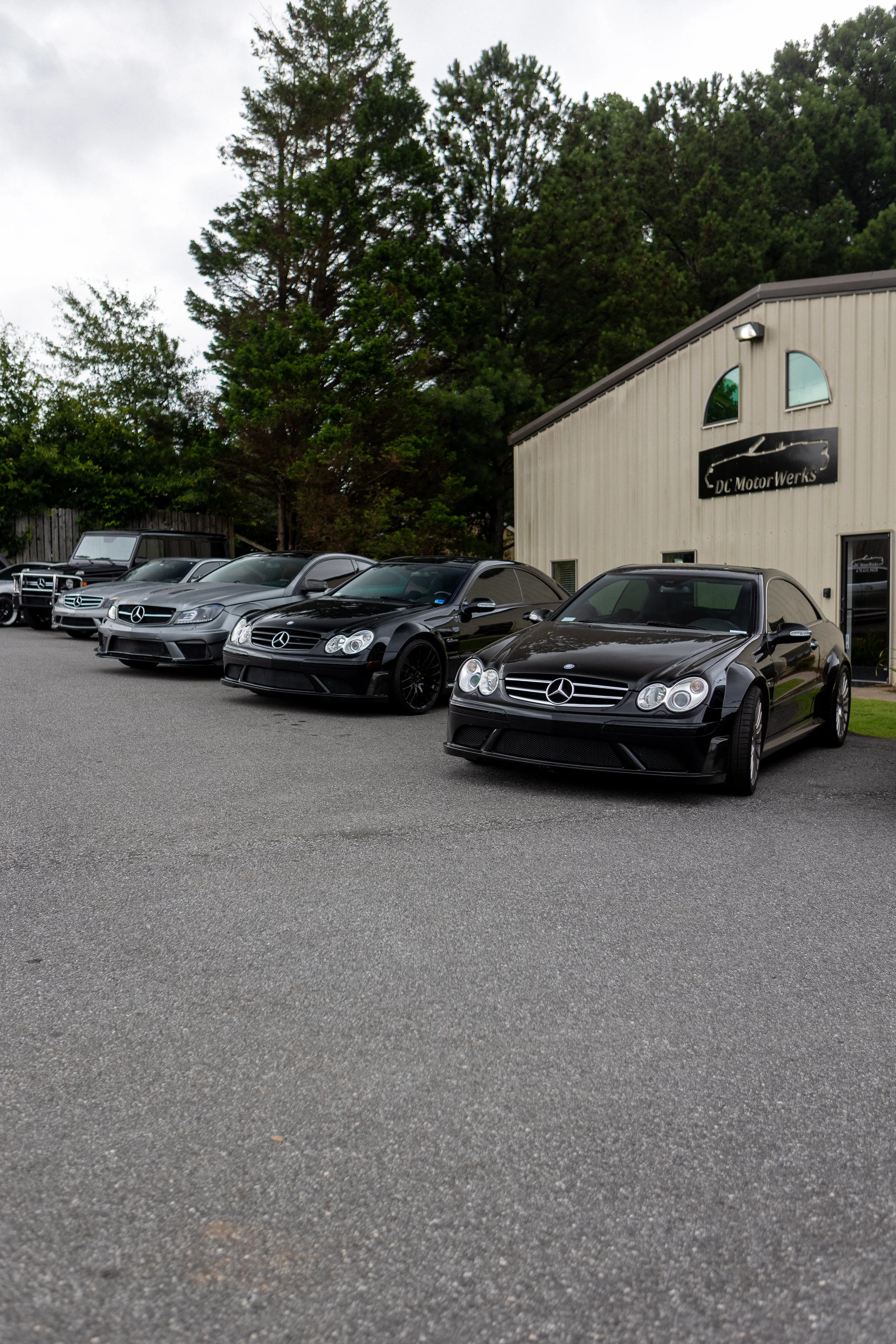 A row of black Mercedes-Benz cars parked outside a beige industrial building labeled 'DC Motor Werks' with green trees in the background.