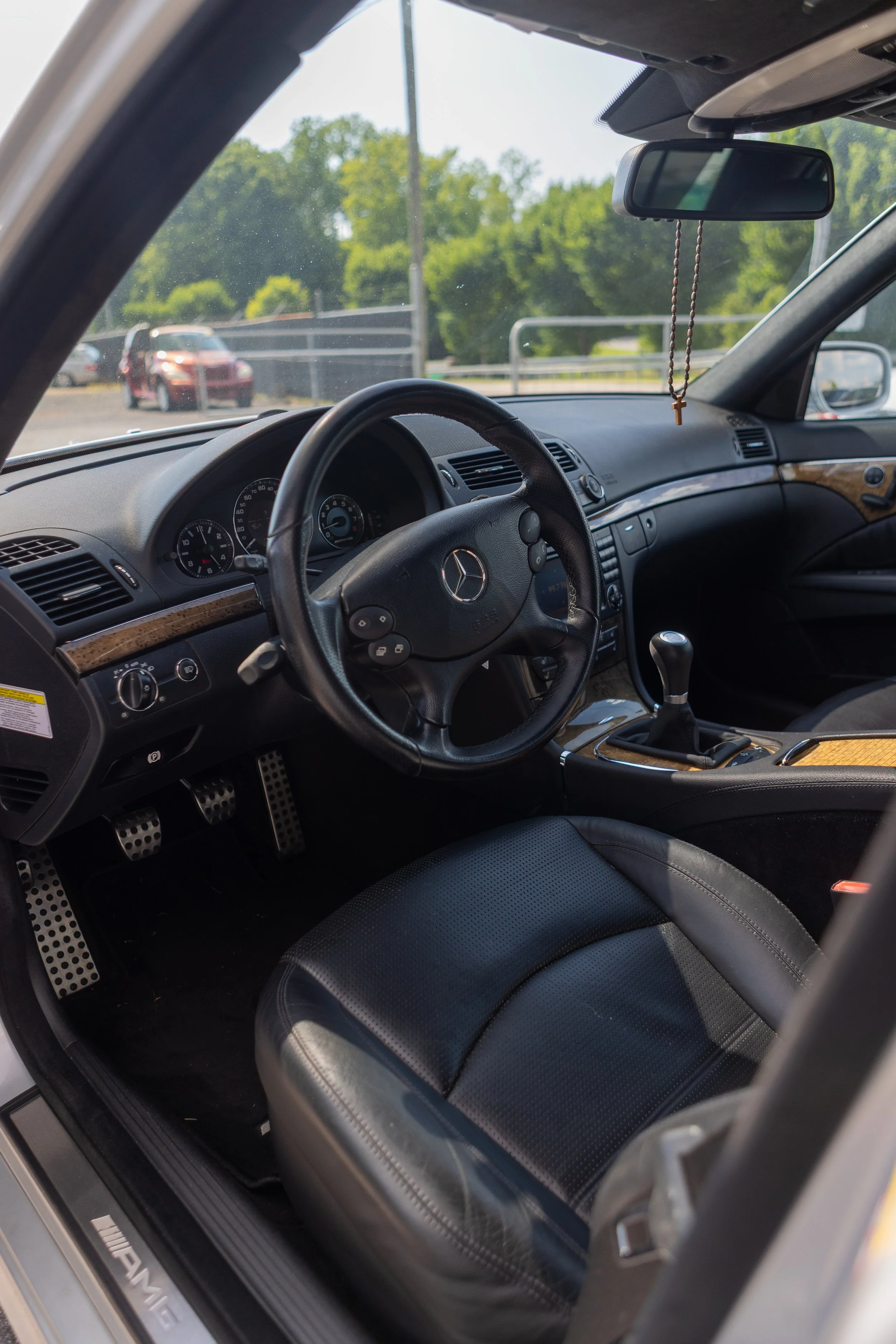 View of the interior of a Mercedes-Benz vehicle, showing the steering wheel, dashboard, gear shifter, black leather seat, and a rosary hanging from the rearview mirror, with trees and parking lot visible outside through the windshield.