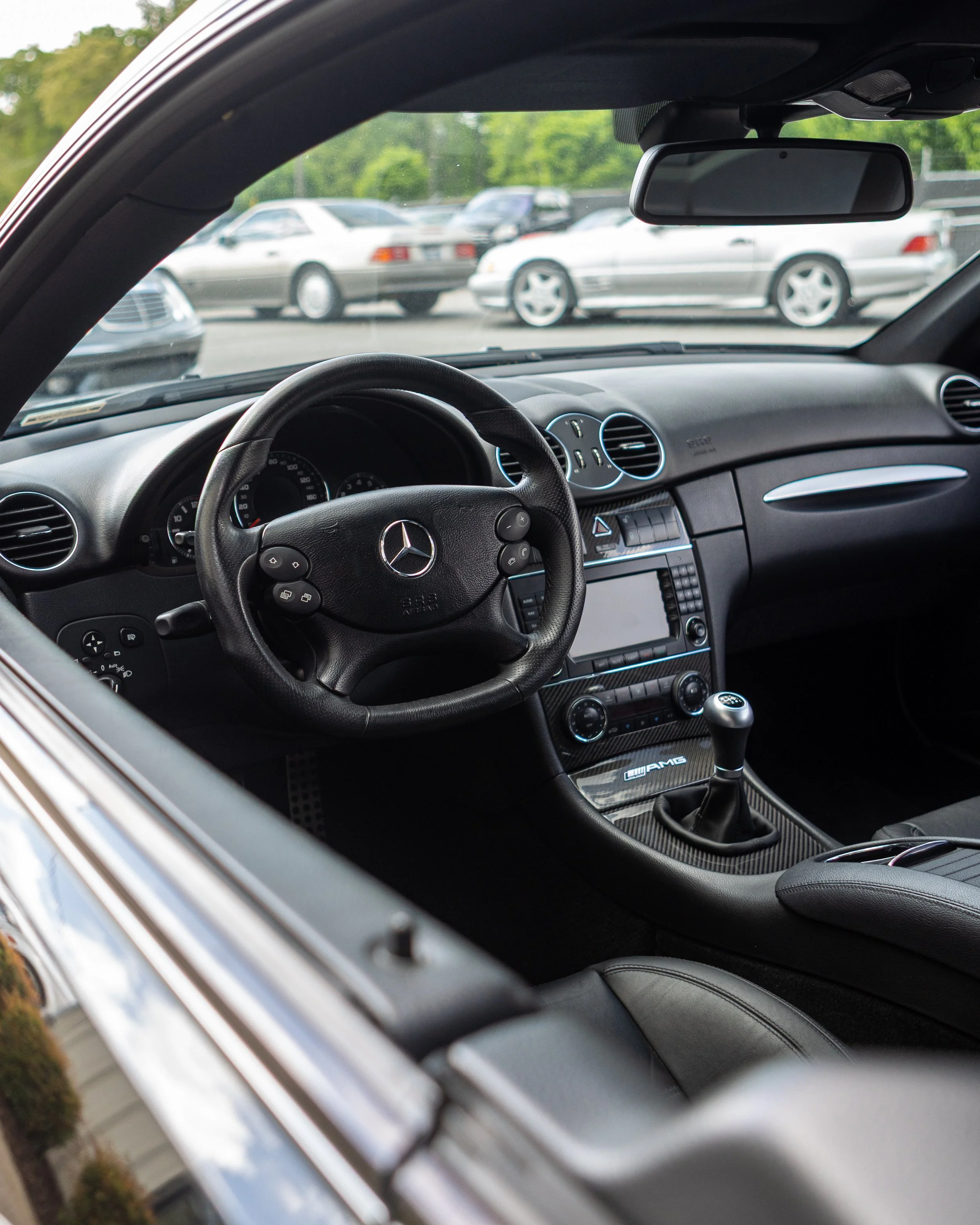 Interior of a Mercedes-Benz car showing the steering wheel, dashboard, gear shift, and rearview mirror with several other cars parked outside visible through the windshield.
