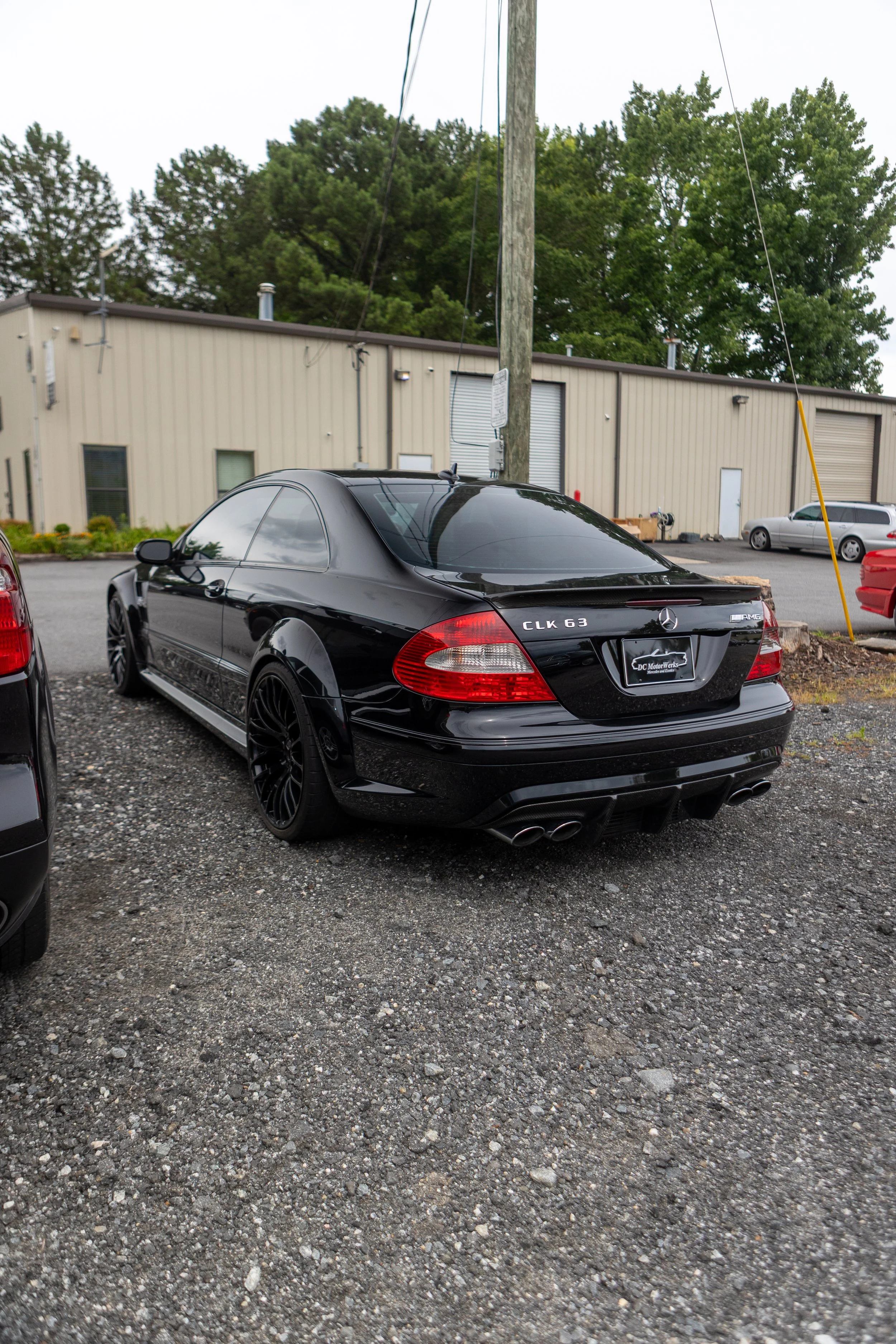 A black Mercedes-Benz CLK 63 AMG parked in a gravel lot, with other cars and a beige industrial building in the background, and trees visible behind the building.