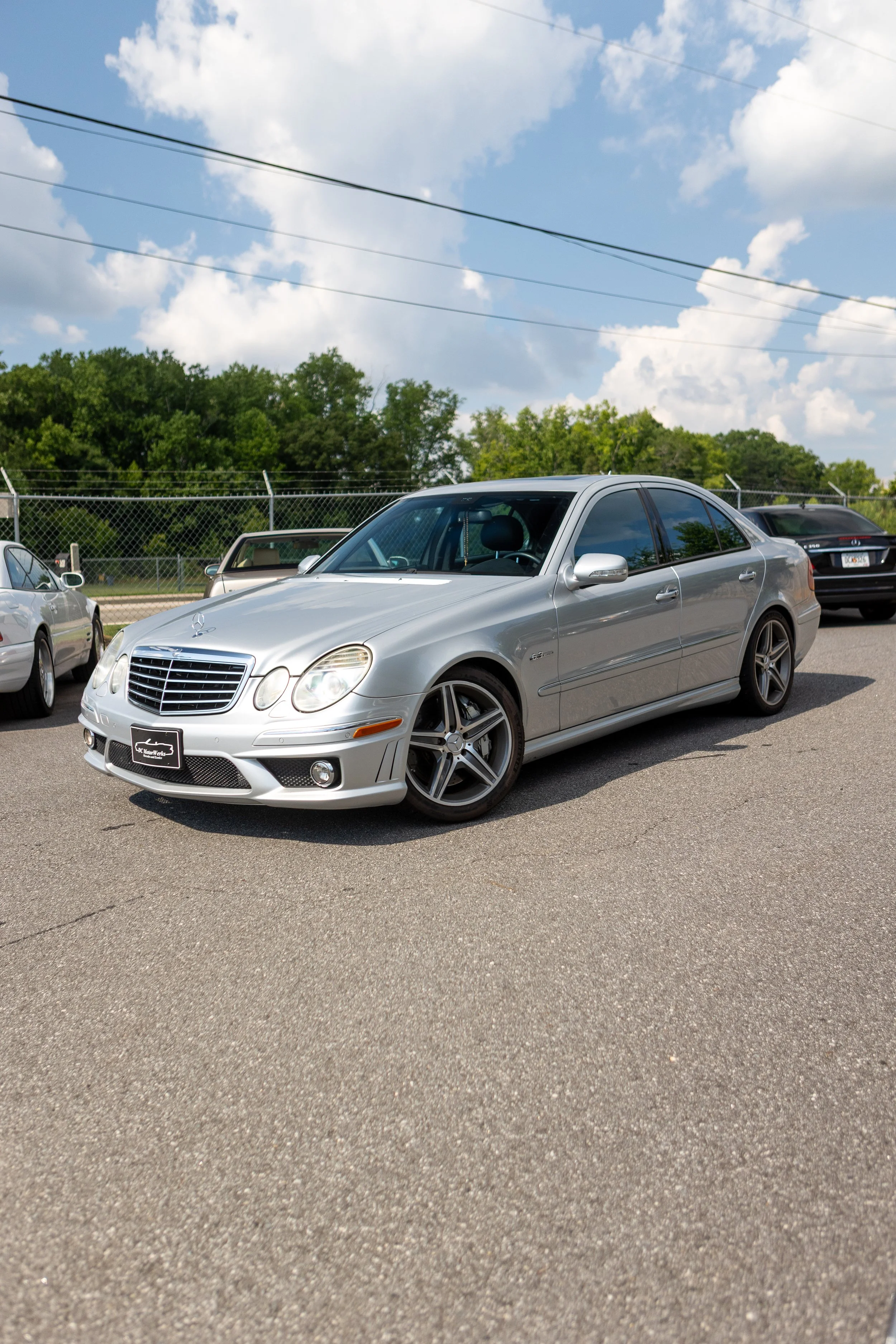 A silver Mercedes-Benz sedan parked in a lot with other cars under a partly cloudy sky, surrounded by a chain-link fence and trees in the background.