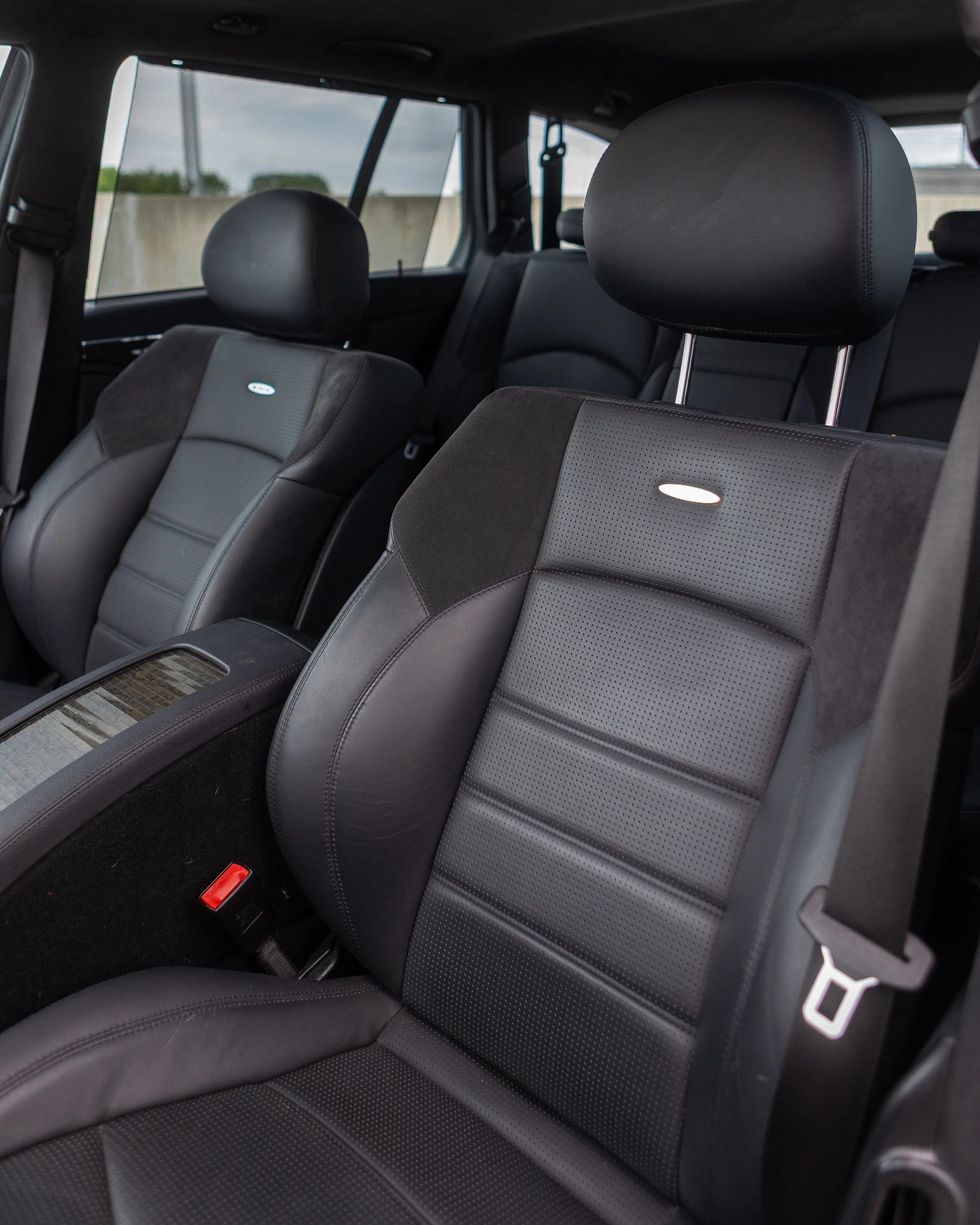 The interior of a vehicle showing black leather seats with perforated patterns and headrests, and a rear window with a view of a concrete wall and cloudy sky.