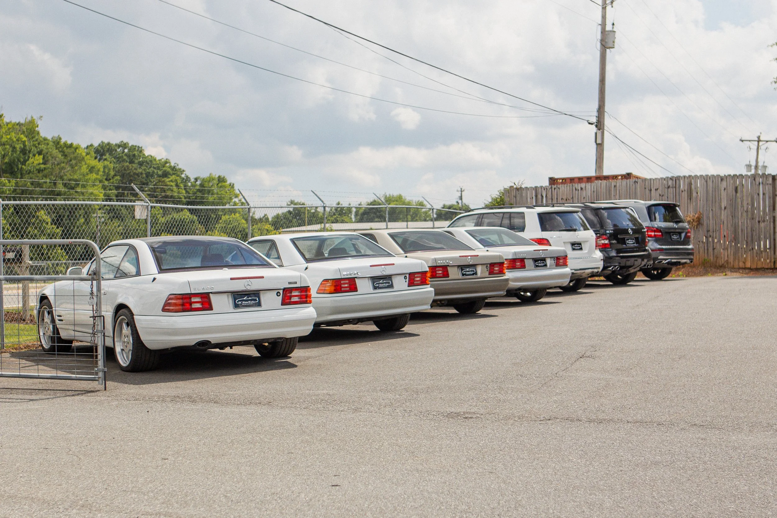 Row of parked Mercedes-Benz cars in parking lot behind a chain-link fence with a wooden fence and utility poles in the background.