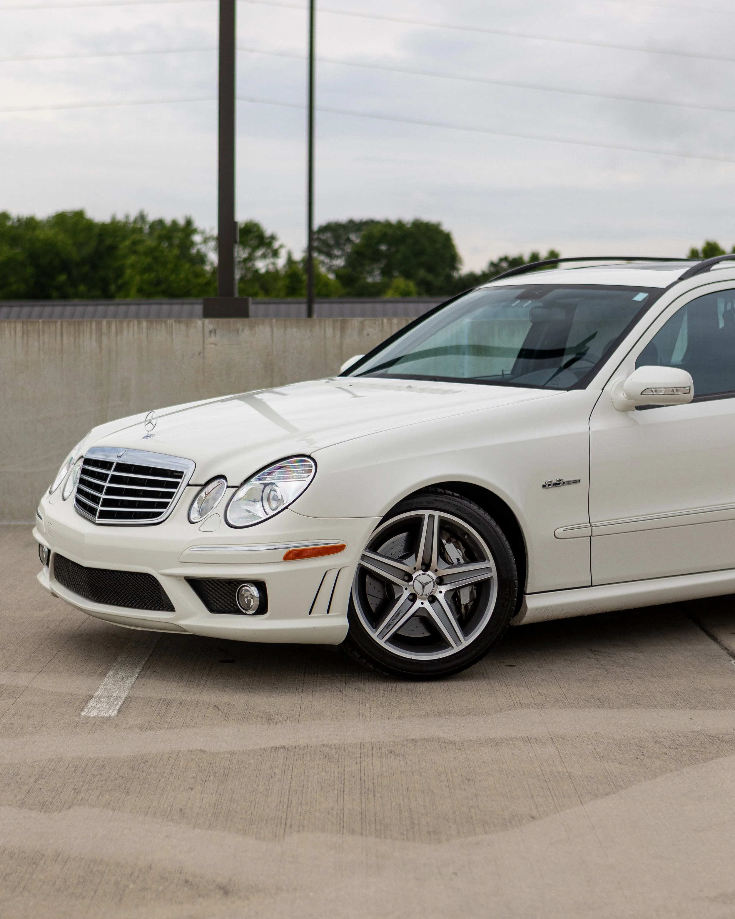 White Mercedes-Benz E-Class parked in a parking lot with green trees and a cloudy sky in the background.