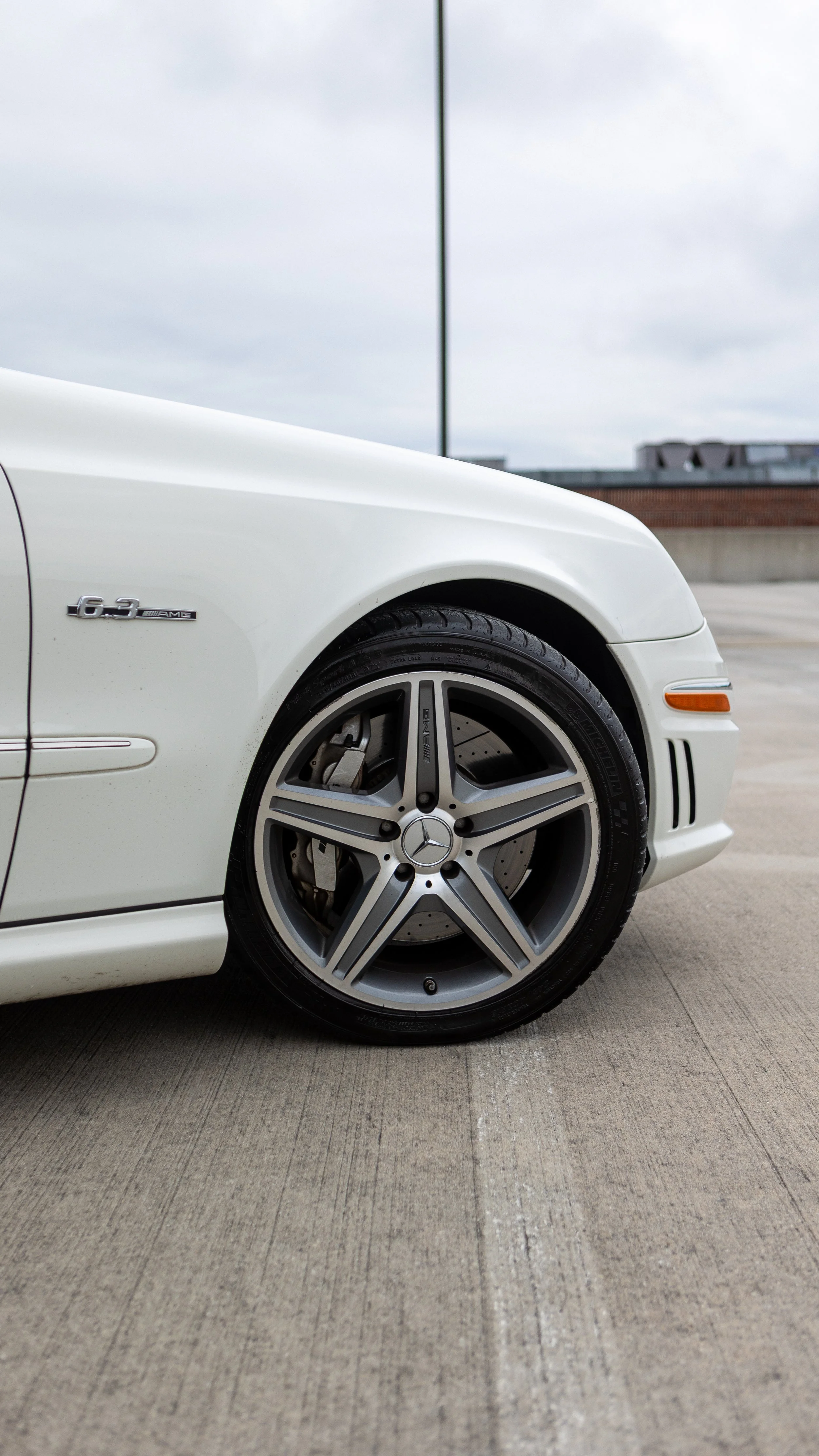 Close-up of the front part of a white Mercedes-Benz car, showing the wheel, part of the front bumper, and a badge that reads '6.3 AMG' on the side.
