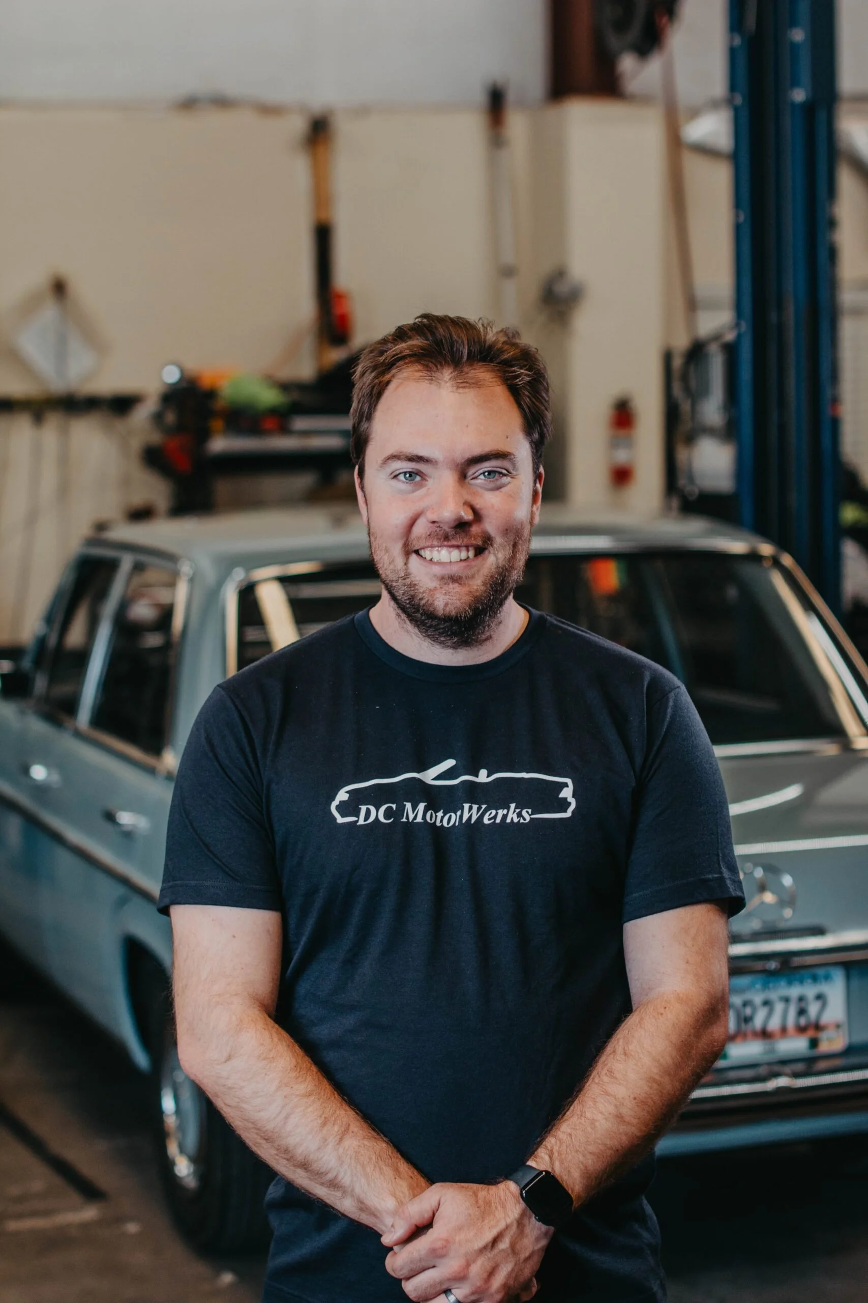 A man with light skin, brown hair, and a beard smiling at the camera inside a garage or workshop. Behind him is a vintage silver car and some tools and equipment on the wall.
