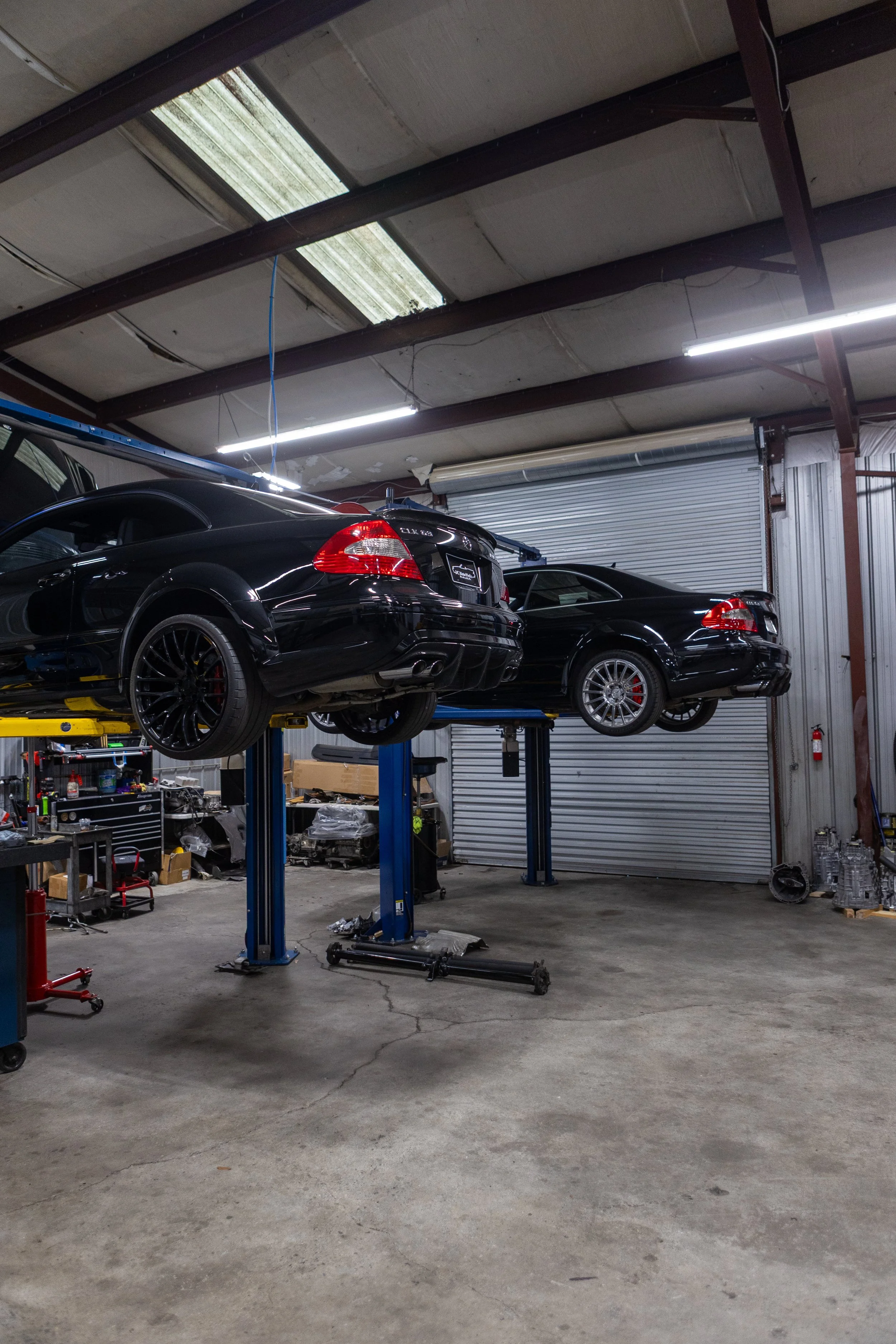 A garage with two black cars lifted on hydraulic lifts, tools and equipment scattered on the concrete floor, a metal roll-up door partially closed, and a ceiling with fluorescent lights and skylights.