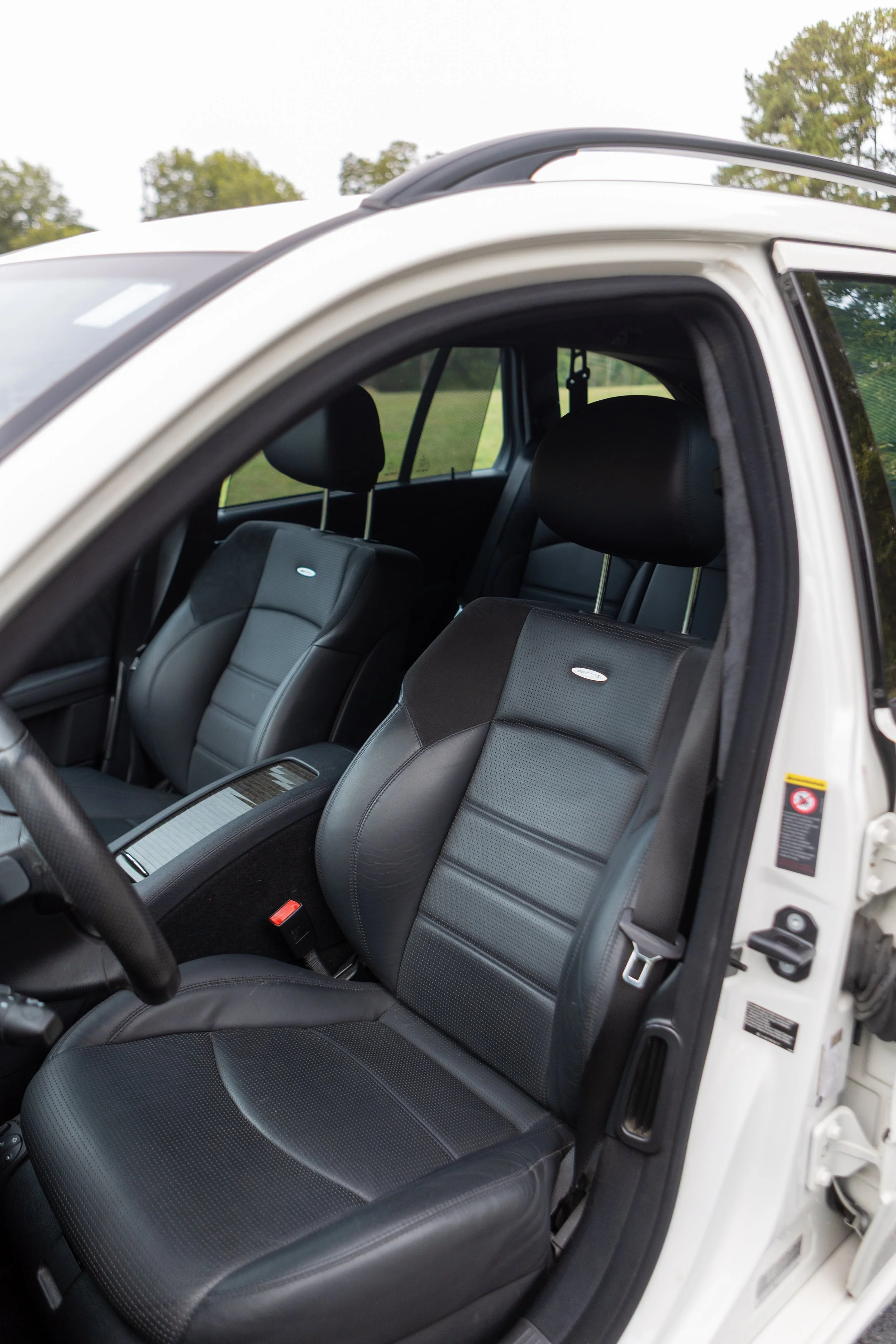 The interior of a vehicle showing black leather seats with perforated patterns and headrests, and a rear window with a view of a concrete wall and cloudy sky.