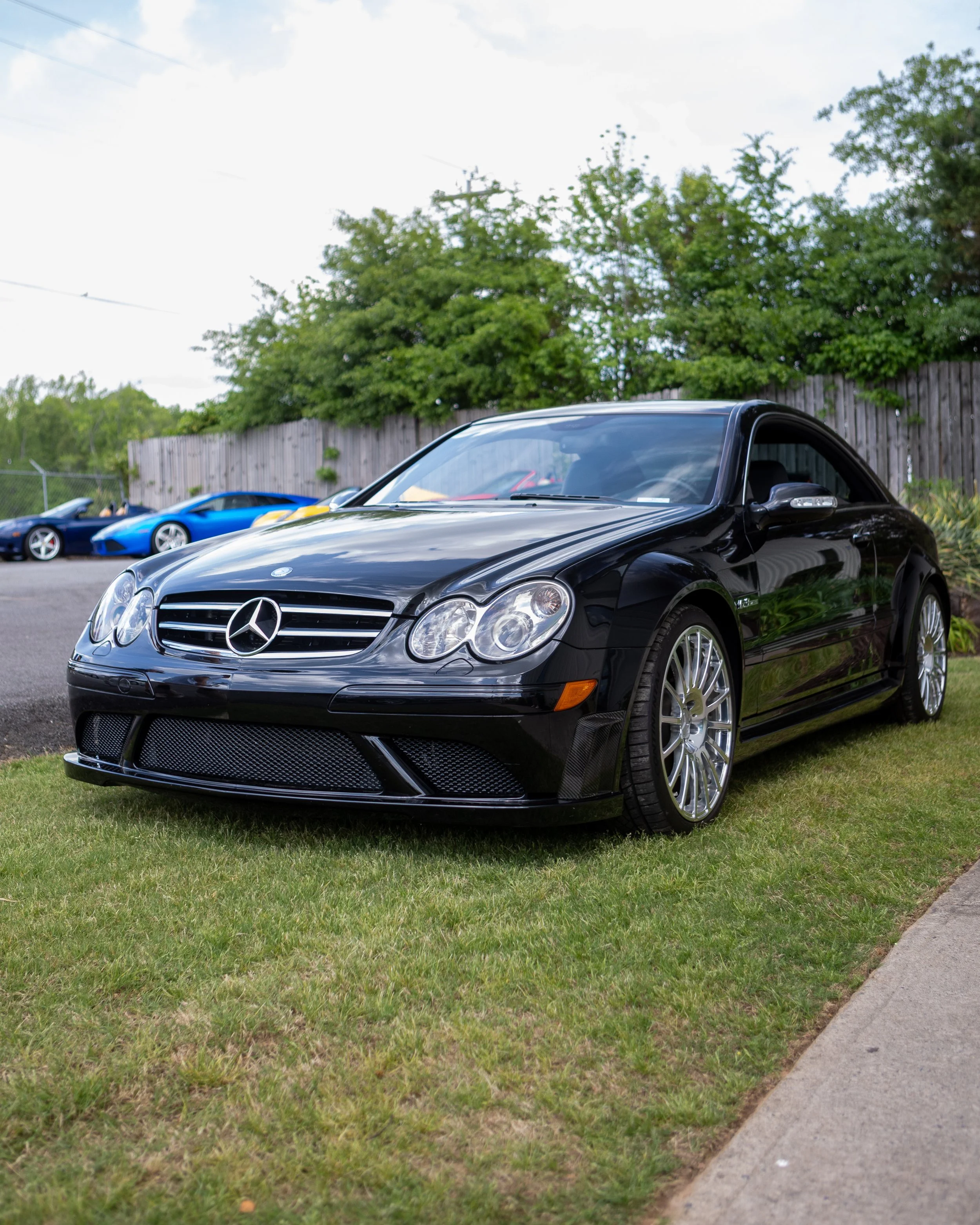 A black Mercedes-Benz coupe parked on a grassy area next to a sidewalk, with a wooden fence and green trees in the background.
