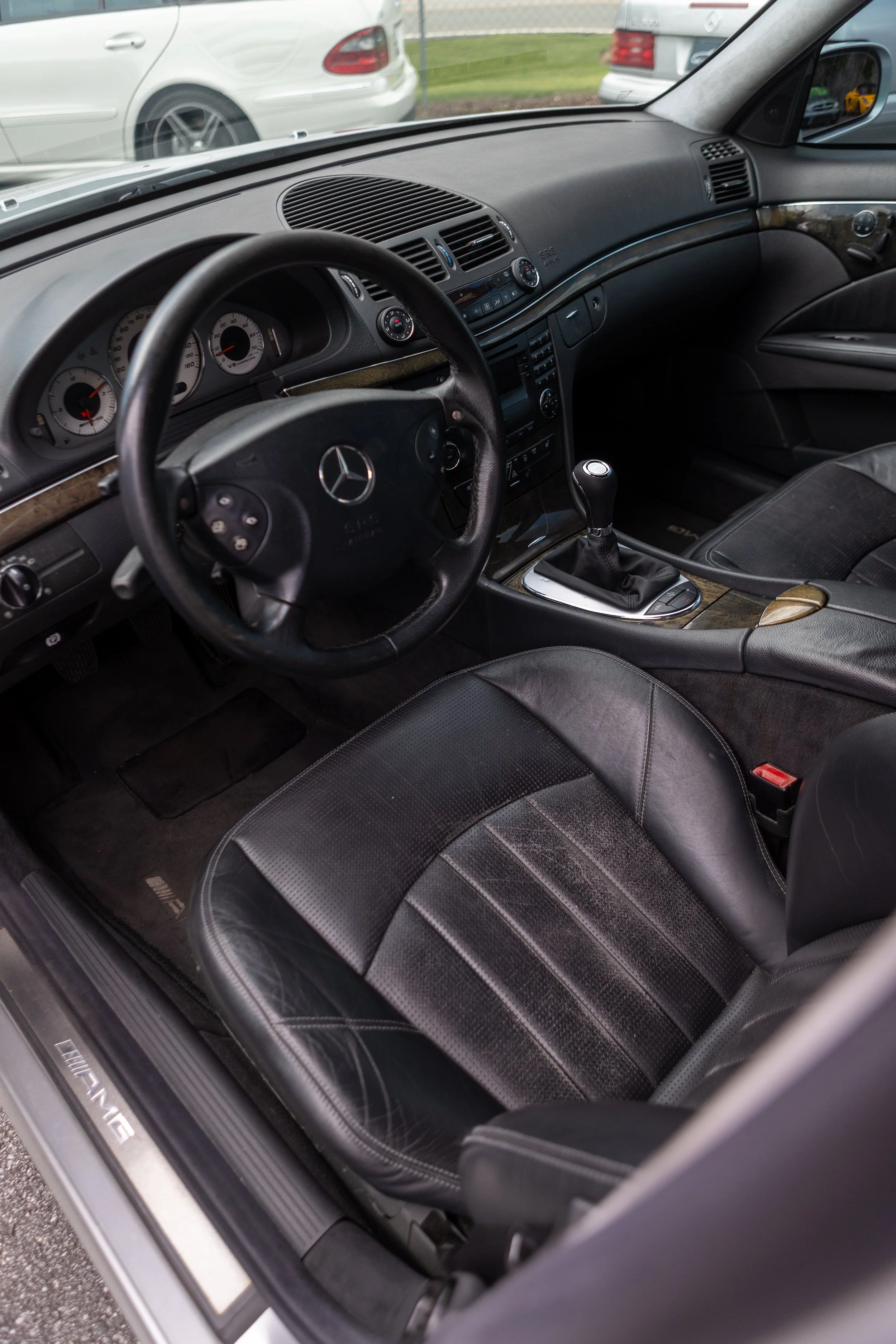 Interior of a Mercedes-Benz vehicle showing the dashboard, steering wheel, gear shift, black leather seats, and driver side door.