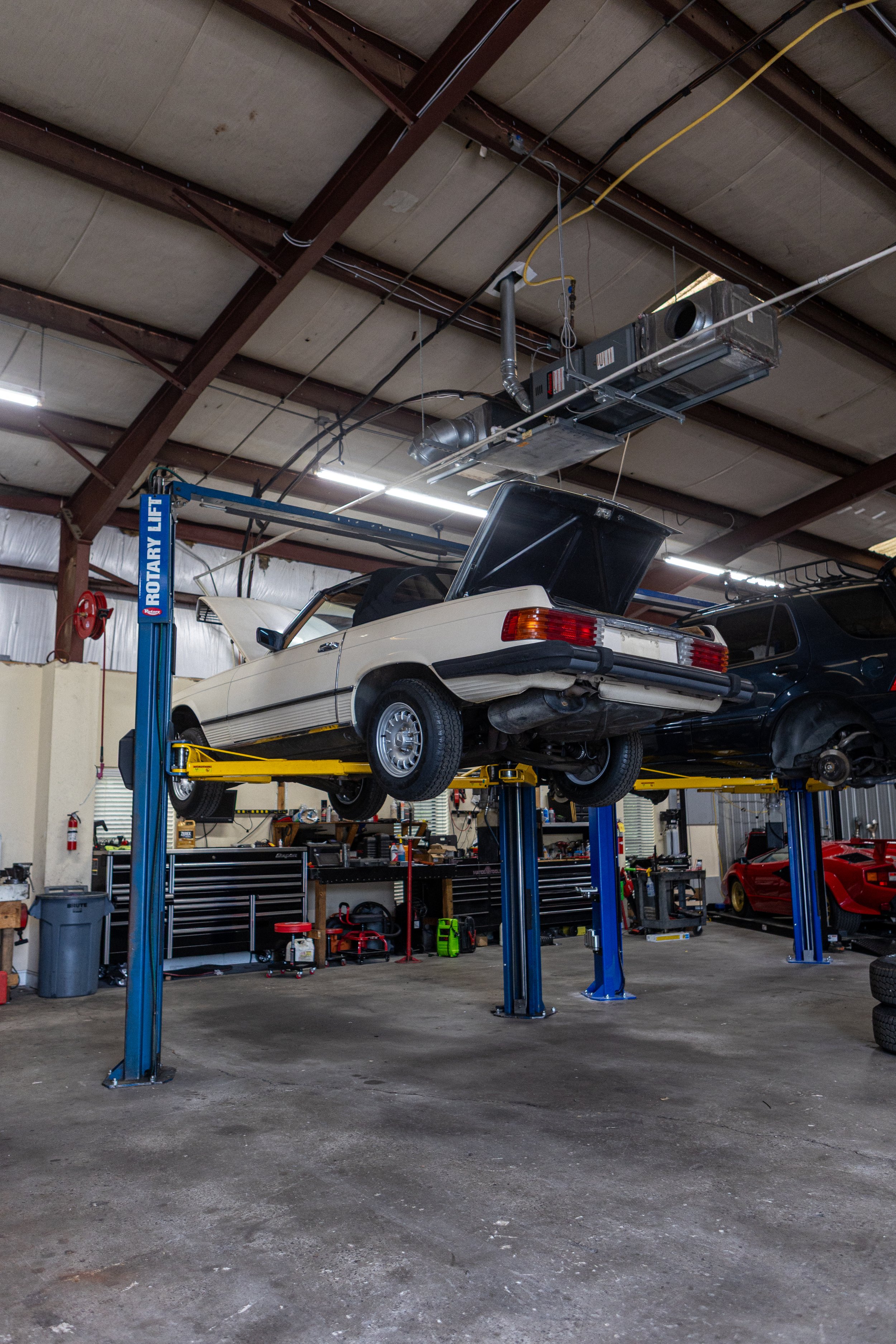 An auto repair garage with a white sports car on a hydraulic lift, and a black vehicle nearby, with various tools and equipment on the workbenches and walls.