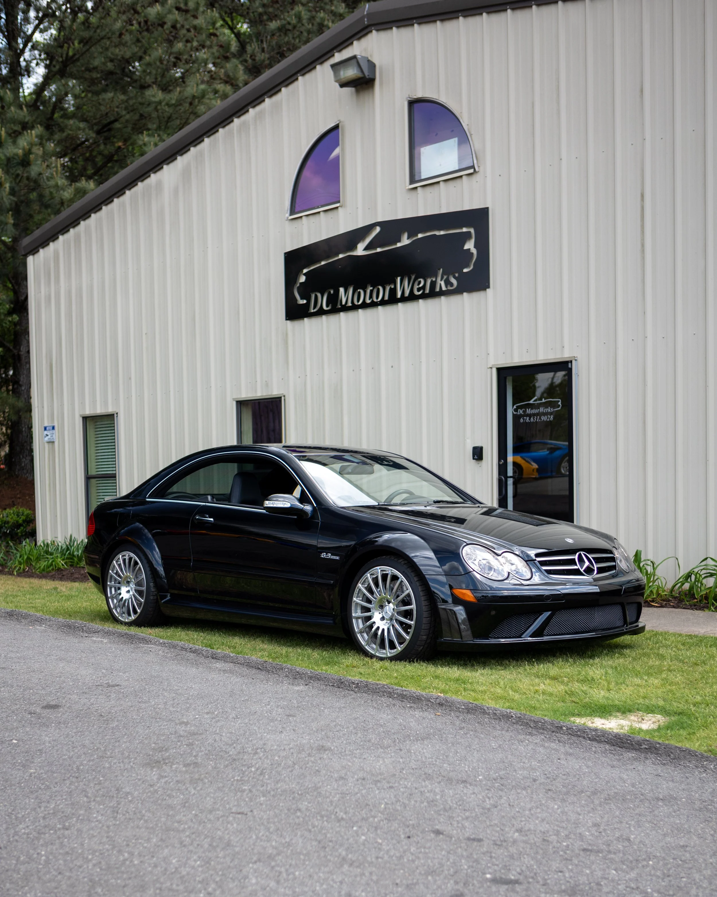 A black Mercedes-Benz sports car parked outside a beige industrial-style building with a sign that reads 'DC MotorWerks.' The building has two arched windows and a door with a small window, surrounded by greenery and trees.