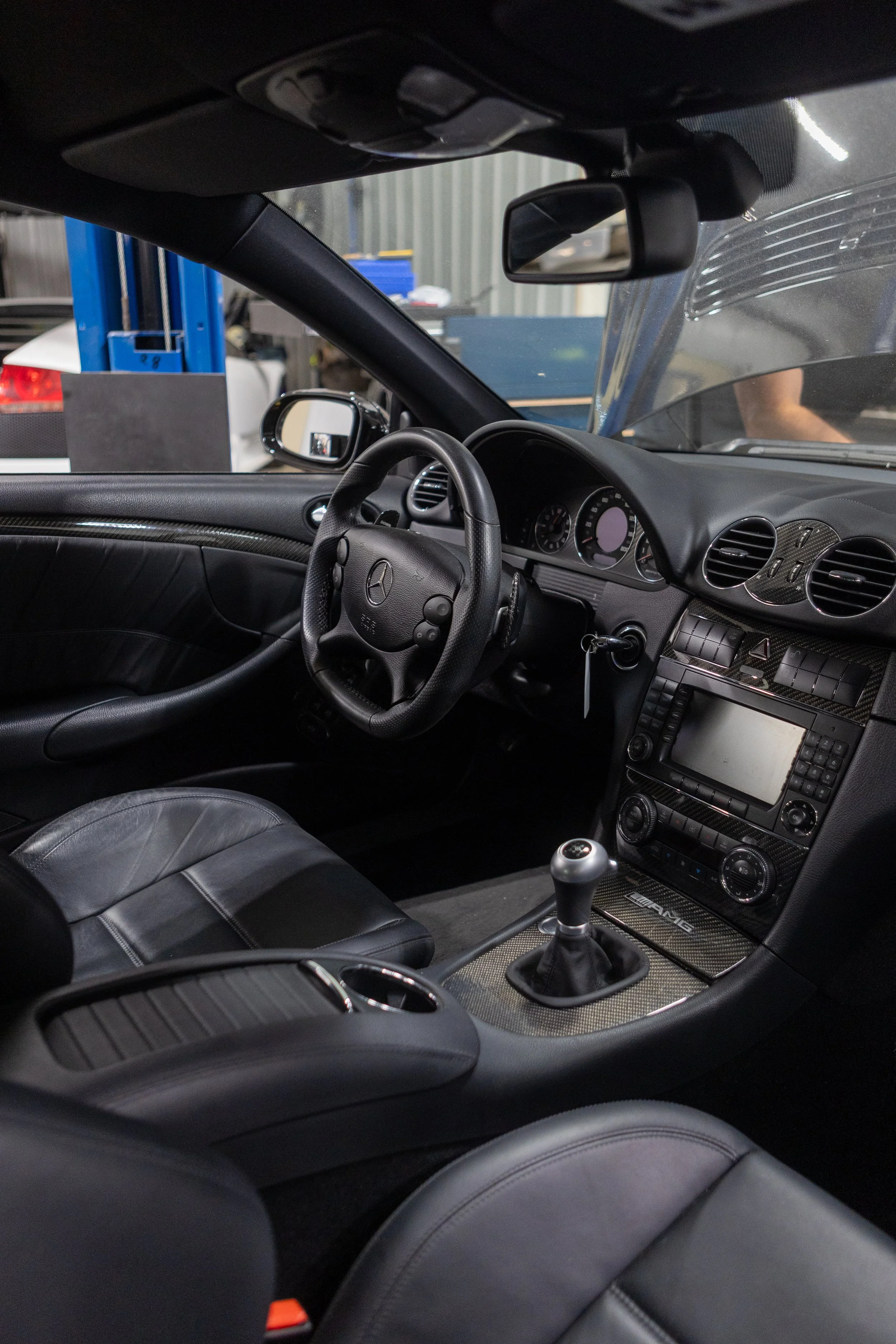 Interior of a black Mercedes-Benz car showing the steering wheel, dashboard, gear shift, and front seats, with a glimpse of the outside through the windshield and side windows, in a garage or workshop setting.