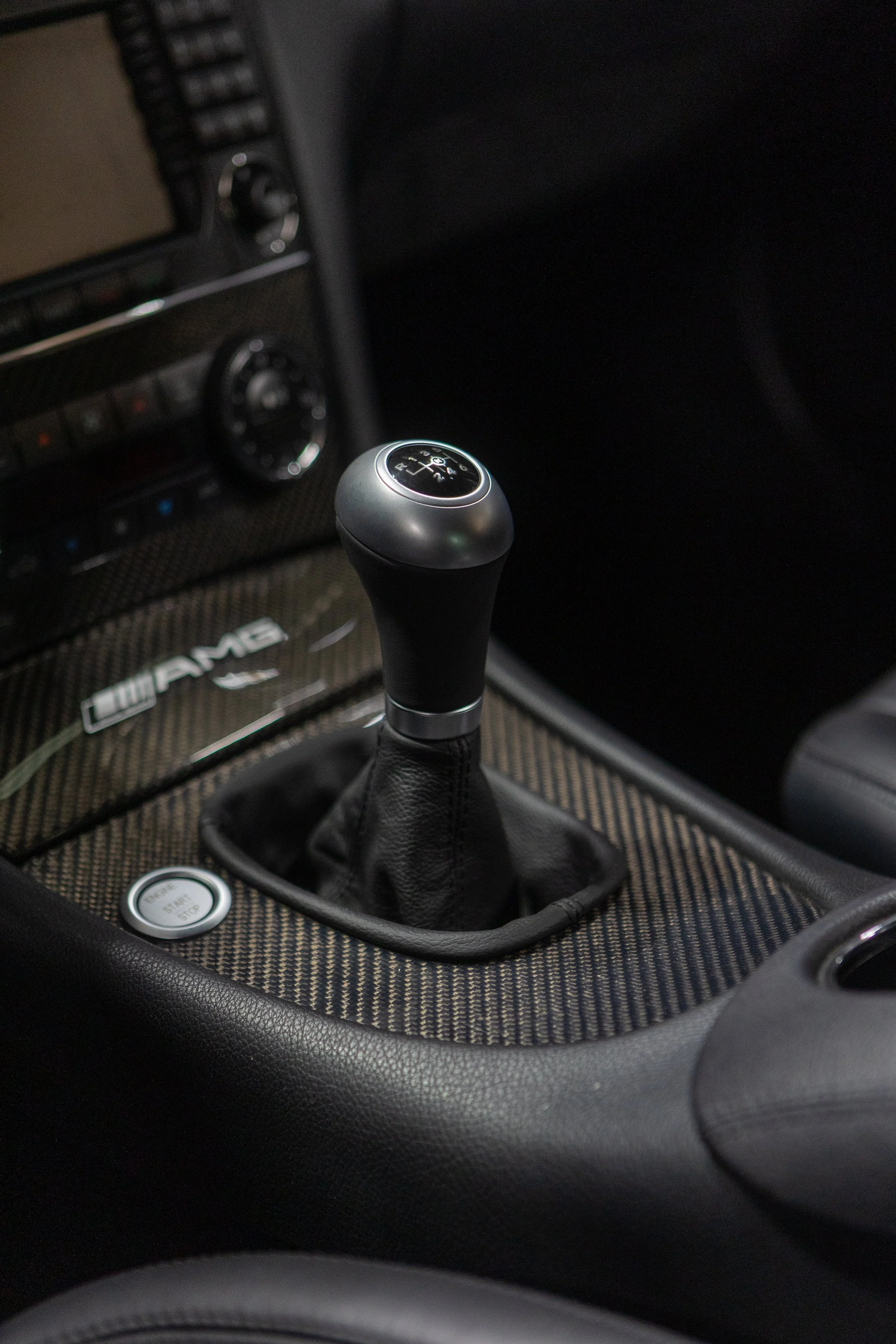 Close-up view of a manual gear shifter in a luxury car interior, with carbon fiber trim and climate control dials in the background.