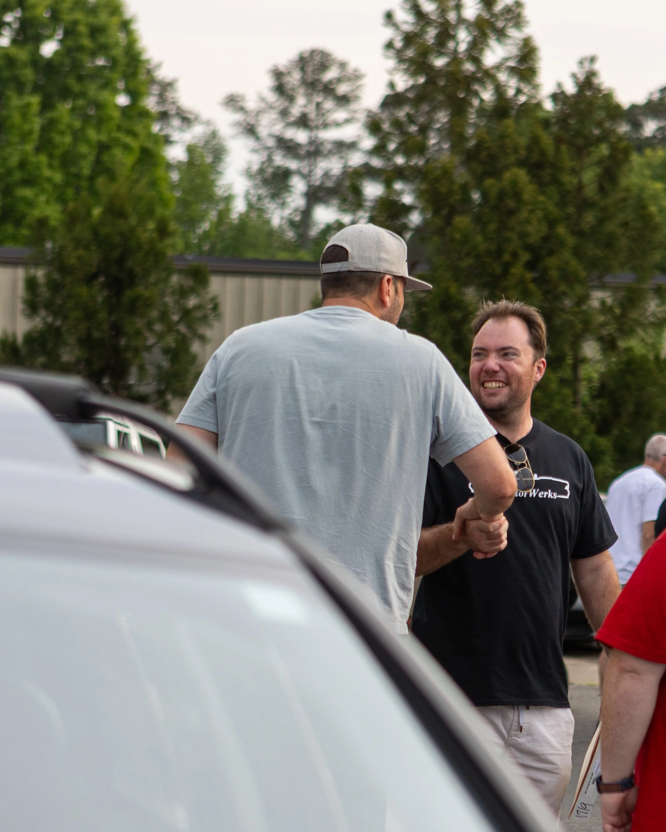 Two men shaking hands outdoors with trees and a building in the background.