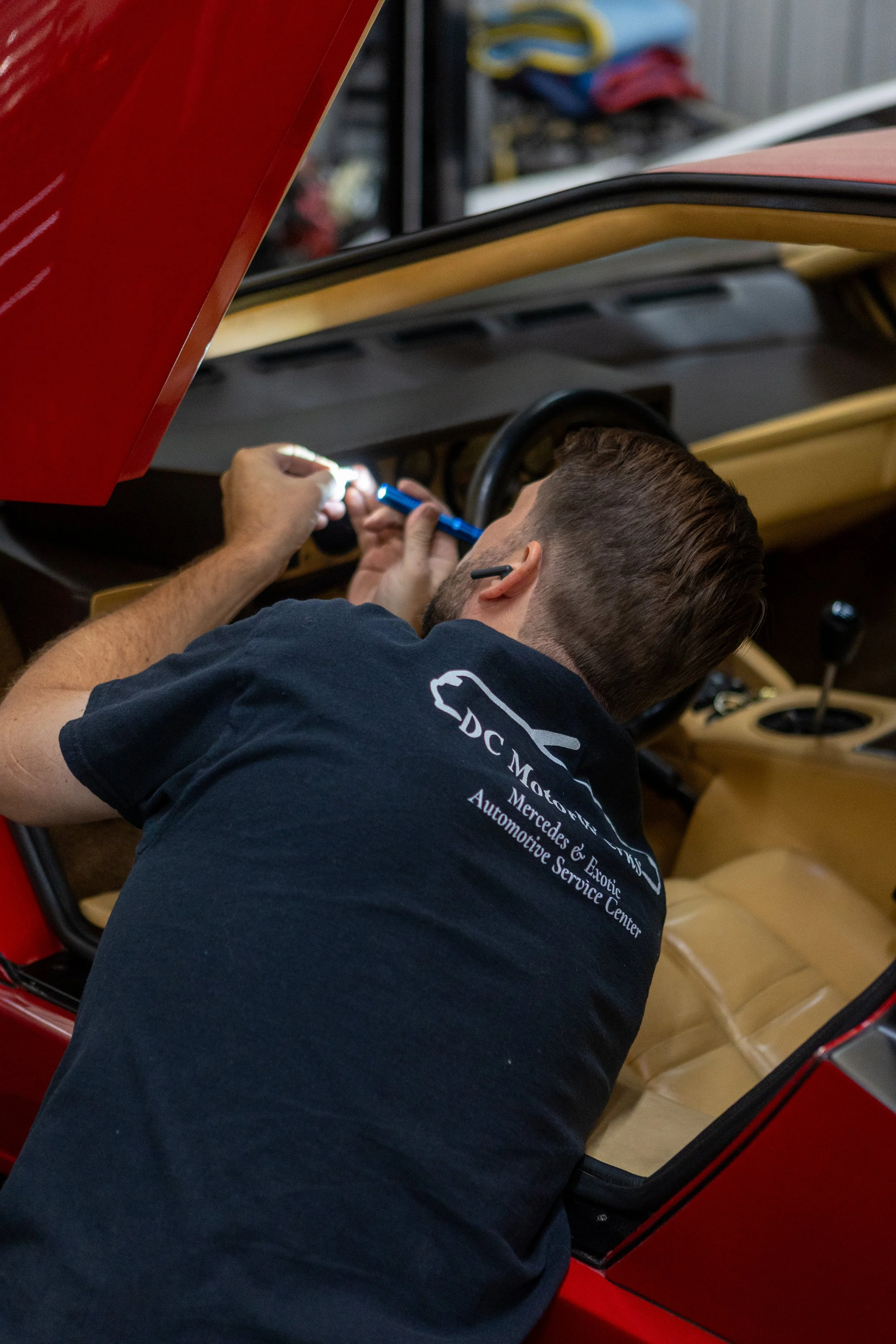 Man working on a red vintage car's interior, focused on the dashboard and steering column, with tools in hand.