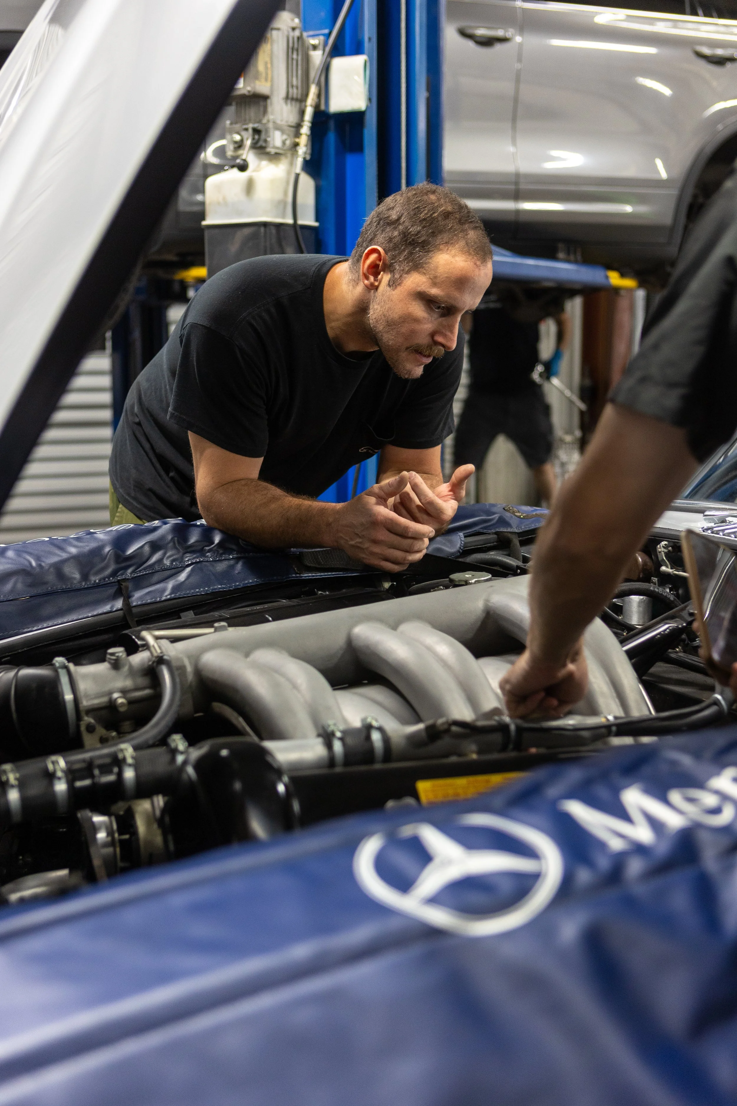 A man working on a car engine in a garage, with a blue hydraulic lift in the background and a gray car elevated above him.