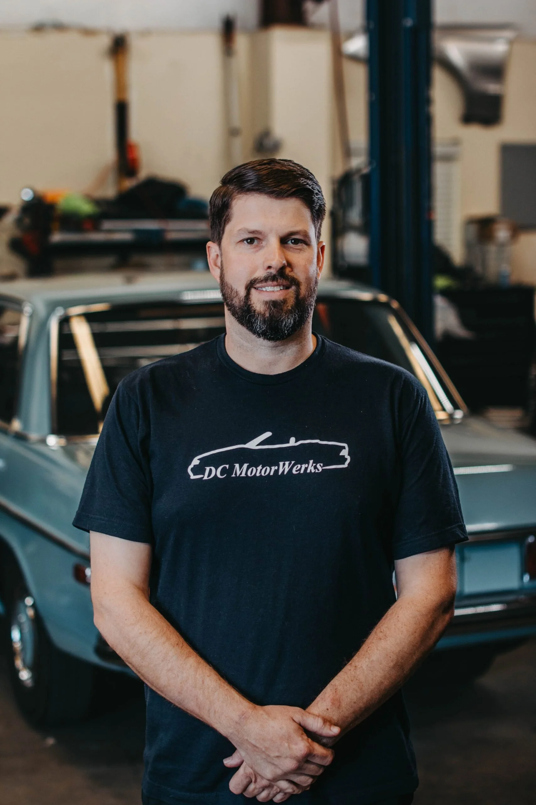 A man with dark hair and a beard standing in front of a vintage car inside a garage or workshop. He is wearing a black T-shirt with the words "DC Motor Werks" on it.