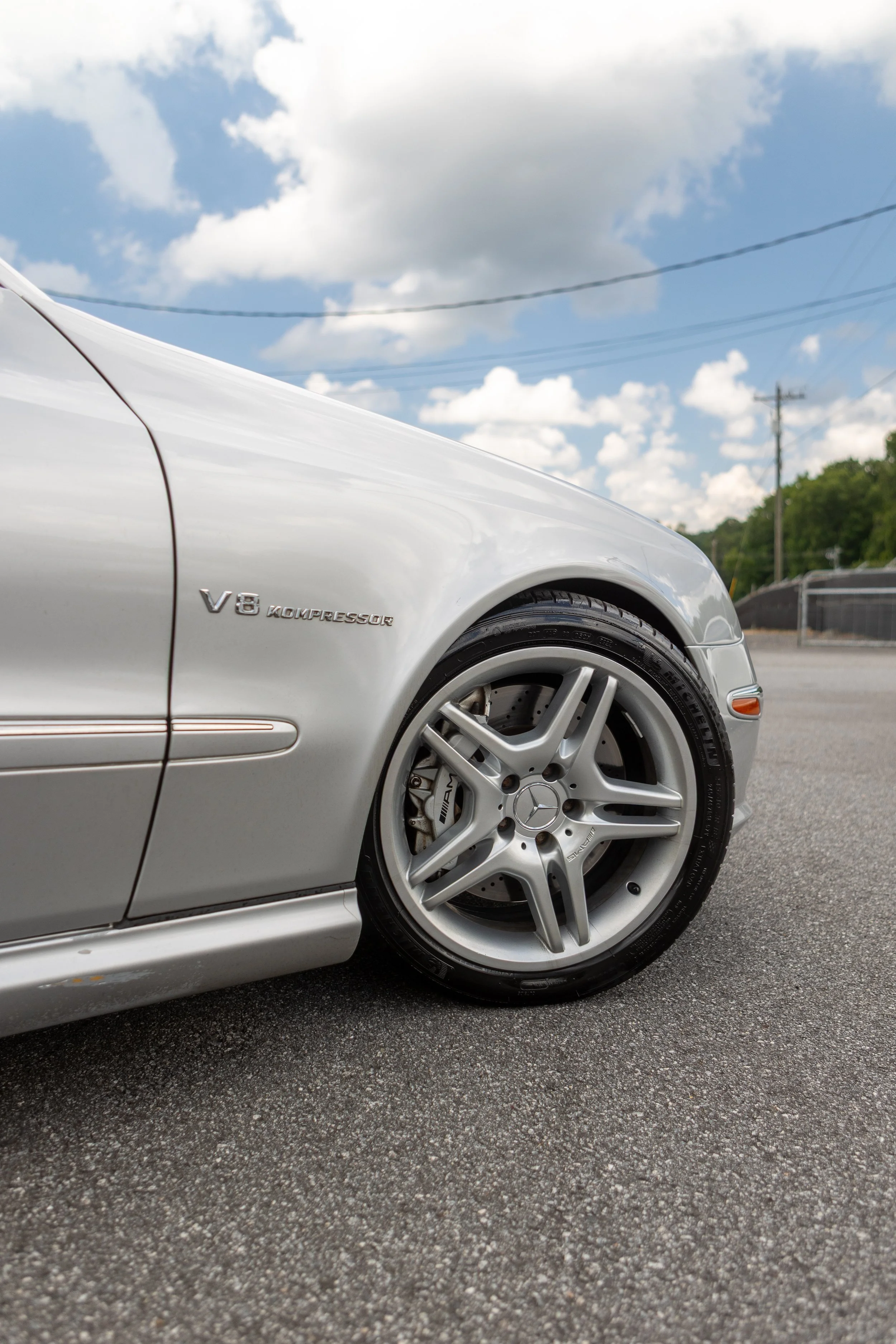 Close-up of the front side of a gray Mercedes-Benz car with a V8 KOMPRESSOR badge and silver alloy wheels, parked on a paved lot with electrical wires and green trees in the background under a partly cloudy sky.