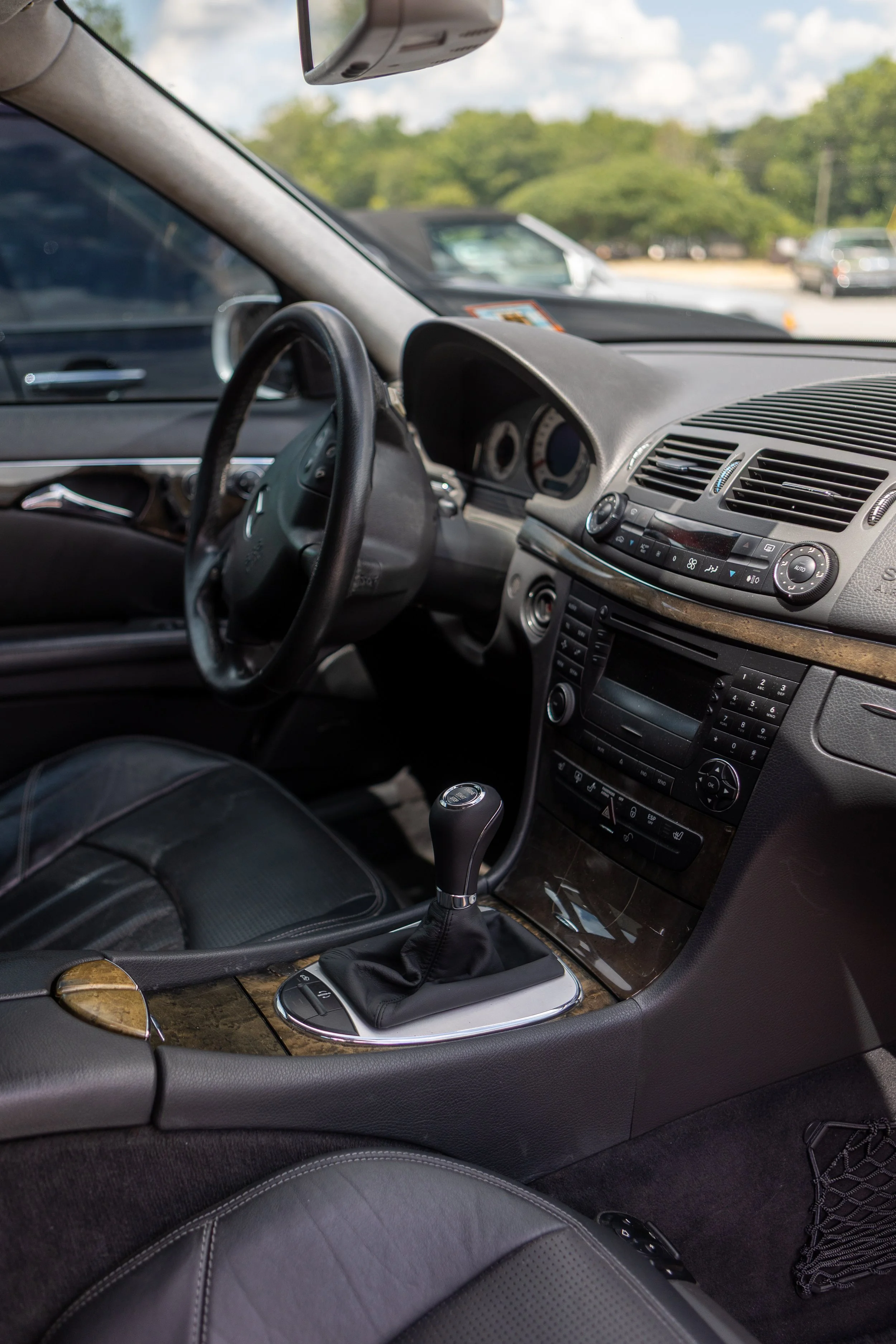 Inside of a car showing the dashboard, steering wheel, gear shift, and part of the front passenger seat. Outside view of a parking lot with trees and several cars.